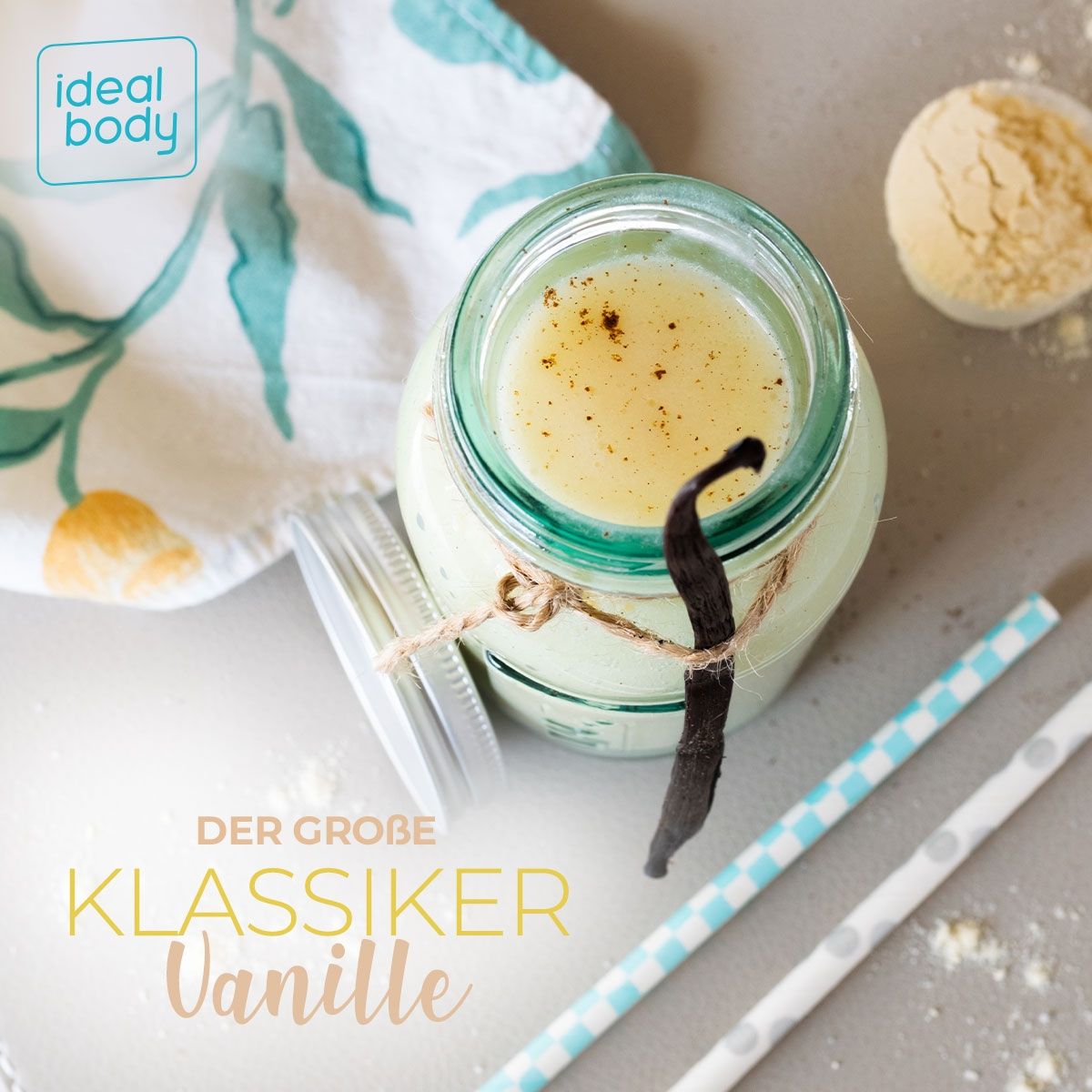 Glas mit Vanille-Proteinshake, Strohhalme, Vanilleschote und Löffel. Aufschrift: "DER GROßE KLASSIKER Vanille". Auf einem Tisch mit Stoff.