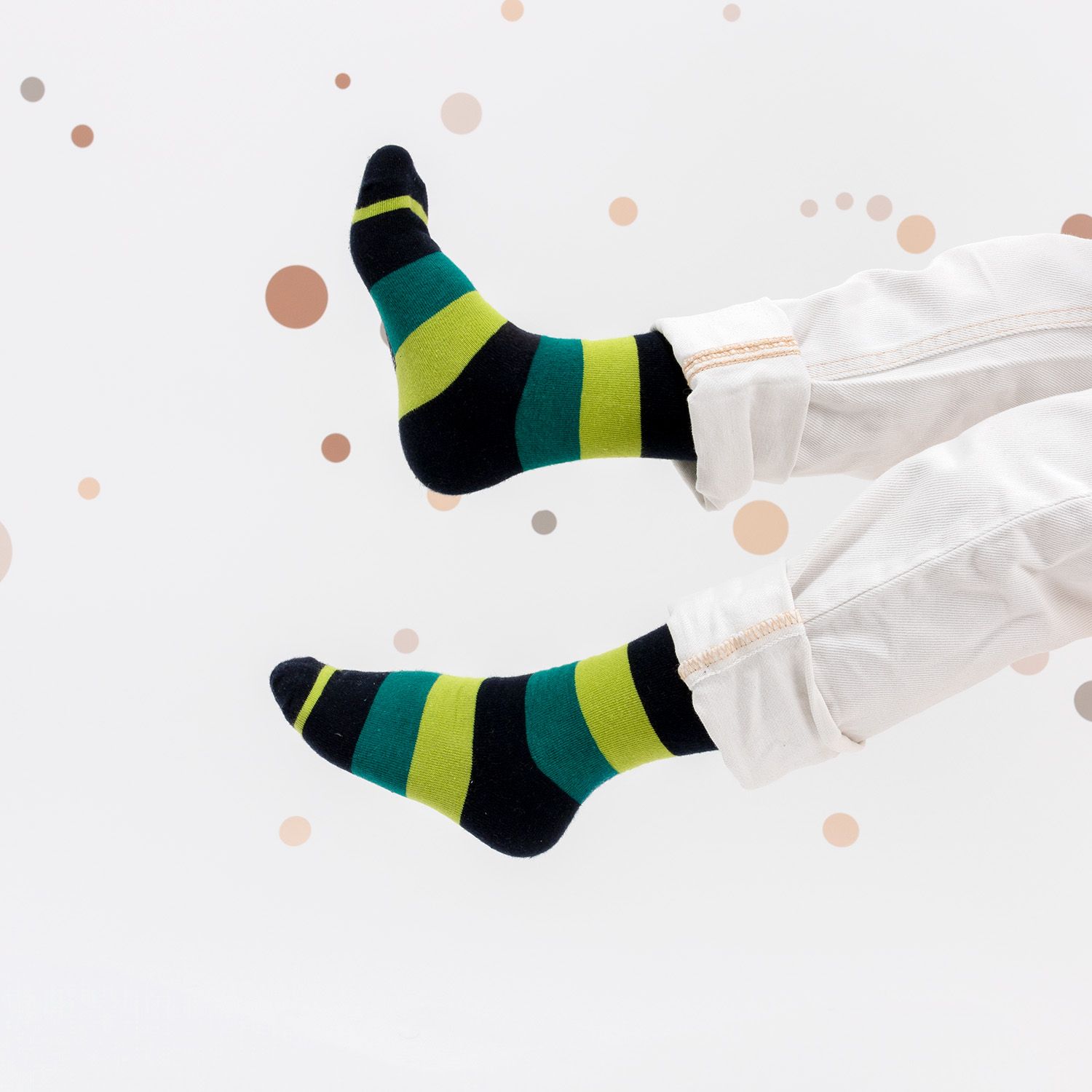 Zwei Füße mit gestreiften Socken in Grün und Schwarz, getragen mit weißer Hose. Hintergrund mit braunen Punkten.