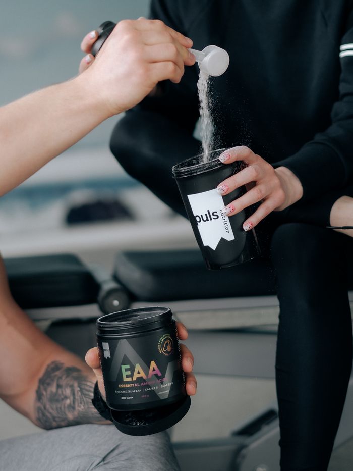 Person hält Dose mit EAA-Aufschrift. Andere Person schüttet Pulver in einen Becher. Schwarze Dose und Becher.