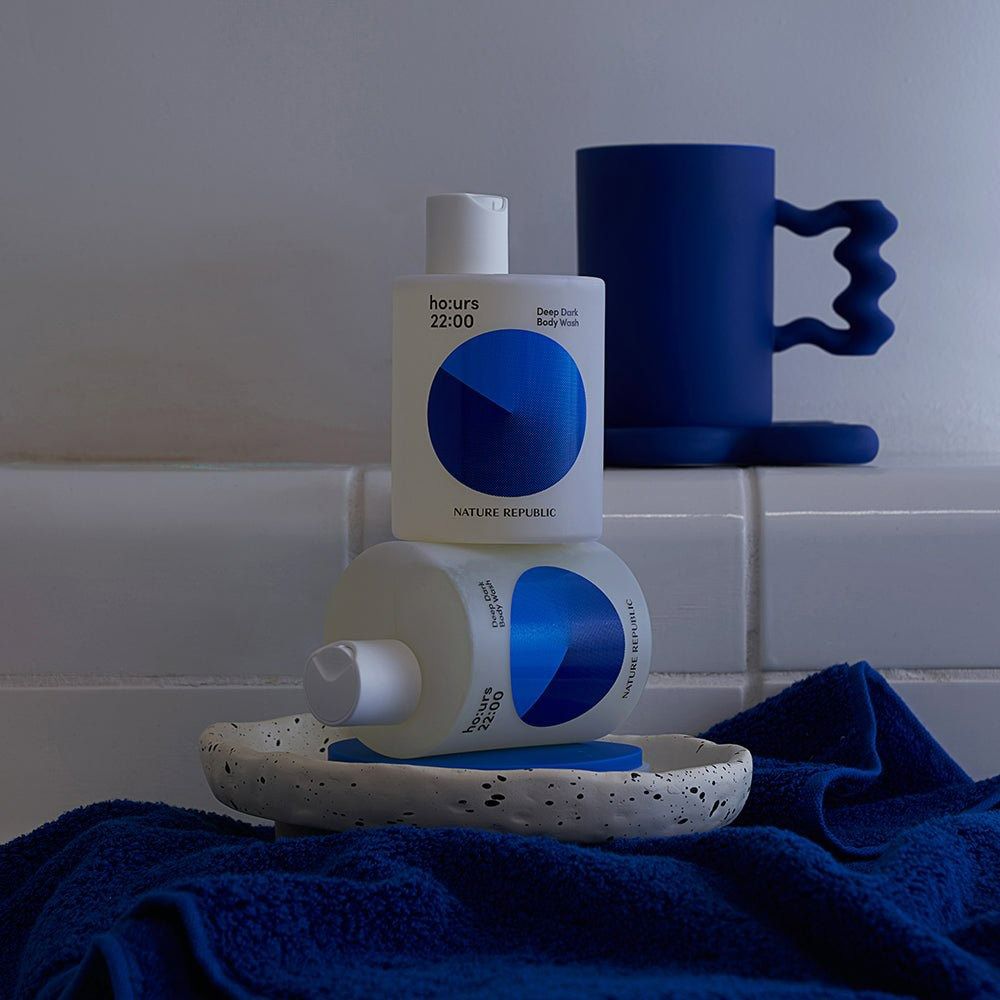 Zwei Duschgel-Flaschen und eine Tasse. Blaue Grafik mit Text: ho:urs 22:00 Deep Dark Body Wash. Marke: NATURE REPUBLIC. Auf einer Seifenschale.