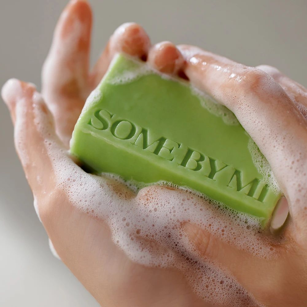 Hände halten grünen Seifenblock mit "SOME BY MI"-Schriftzug, umgeben von Schaum.