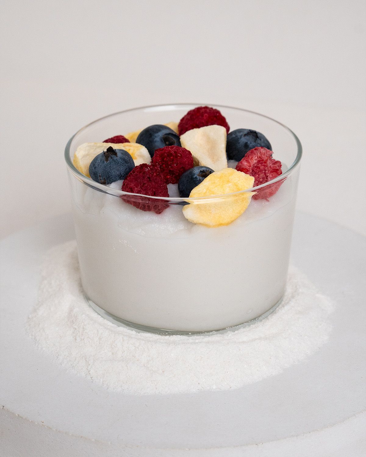 Glas mit weißem Brei, belegt mit Blaubeeren, Himbeeren und Bananenscheiben. Auf weißem Teller.