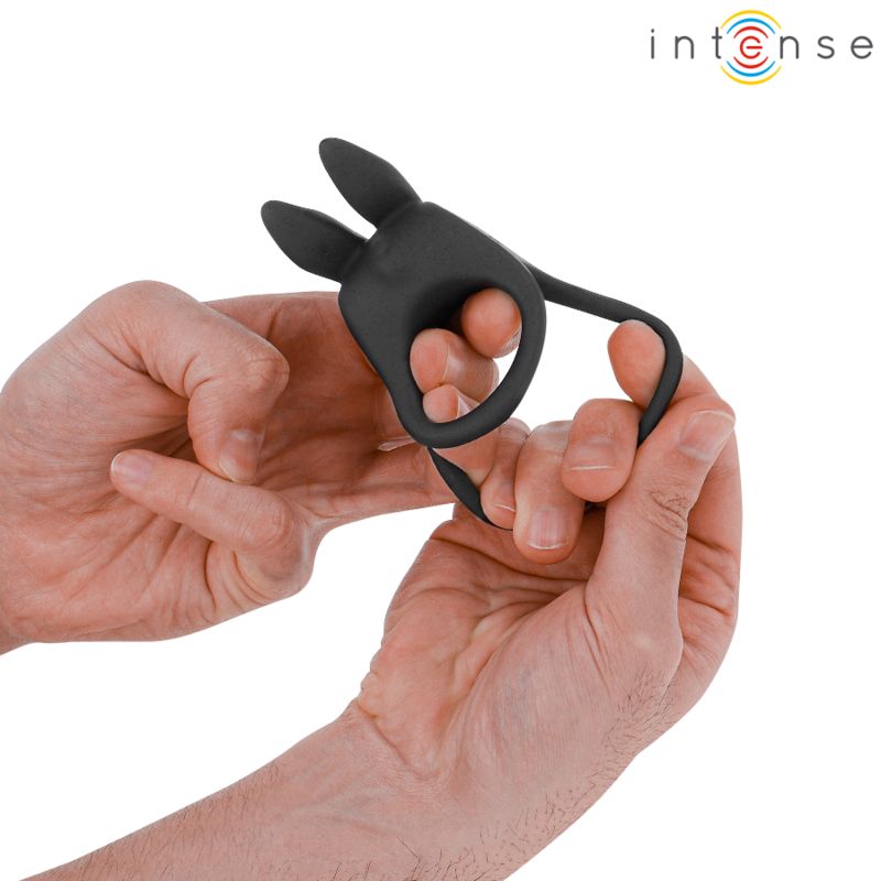 Person hält schwarzen Doppel-Penisring. Ring mit zwei Öffnungen und einem blauen Knopf. Ring wird mit den Händen gehalten.