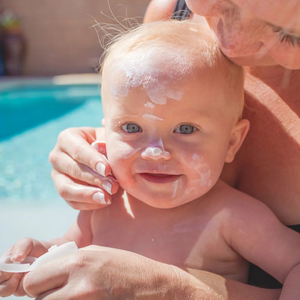 Ein Baby mit Sonnencreme im Gesicht, gehalten von einer Person am Pool.