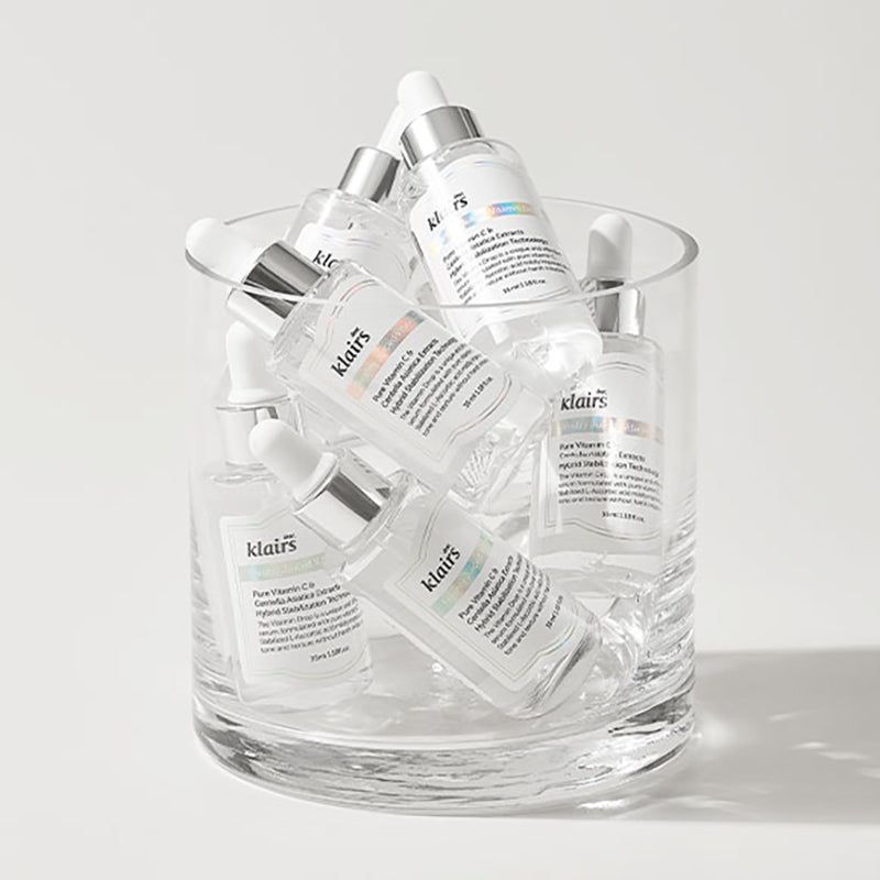 Mehrere Klairs Serumflaschen in einem Glasbehälter. Klares Glas, silberne Deckel, weiße Etiketten.