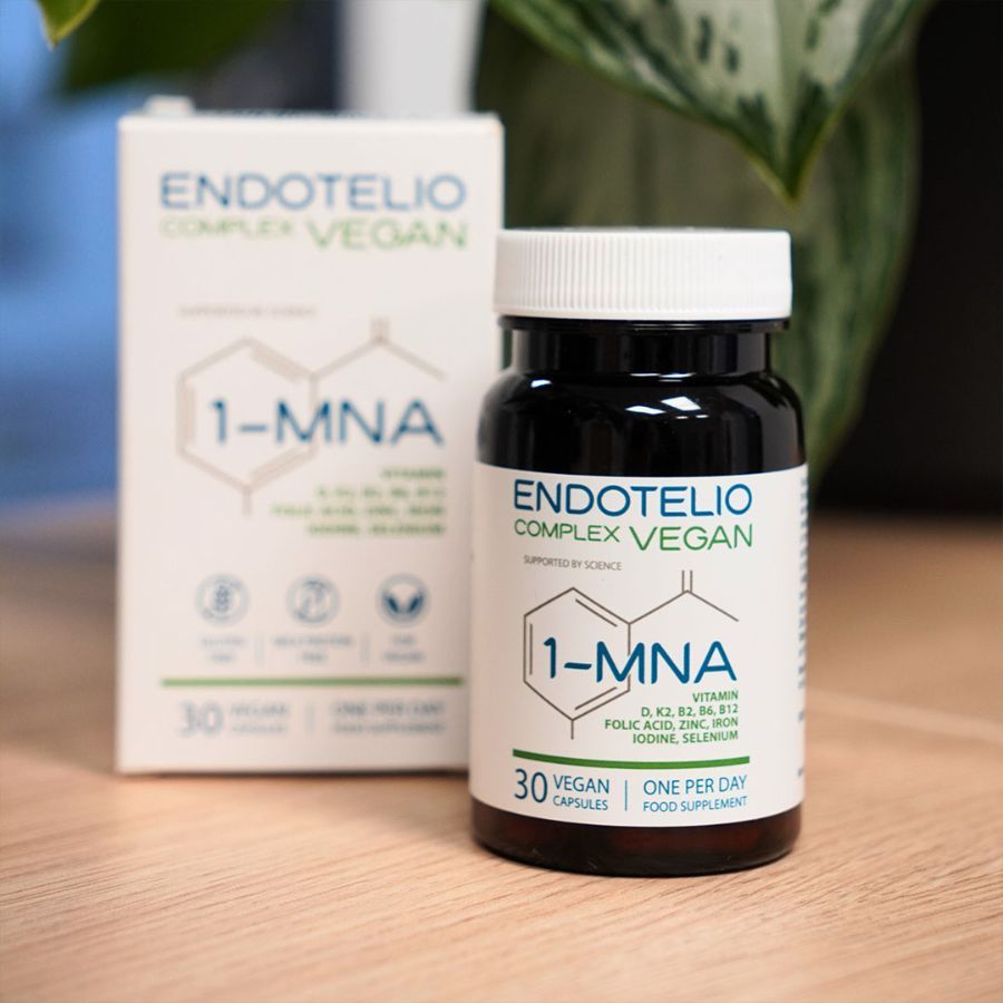 Braune Flasche und Schachtel mit Produktinformationen. Aufschrift: Endotelio Complex Vegan, 1-MNA, 30 Kapseln. Steht auf einem Tisch, im Hintergrund Pflanzen.