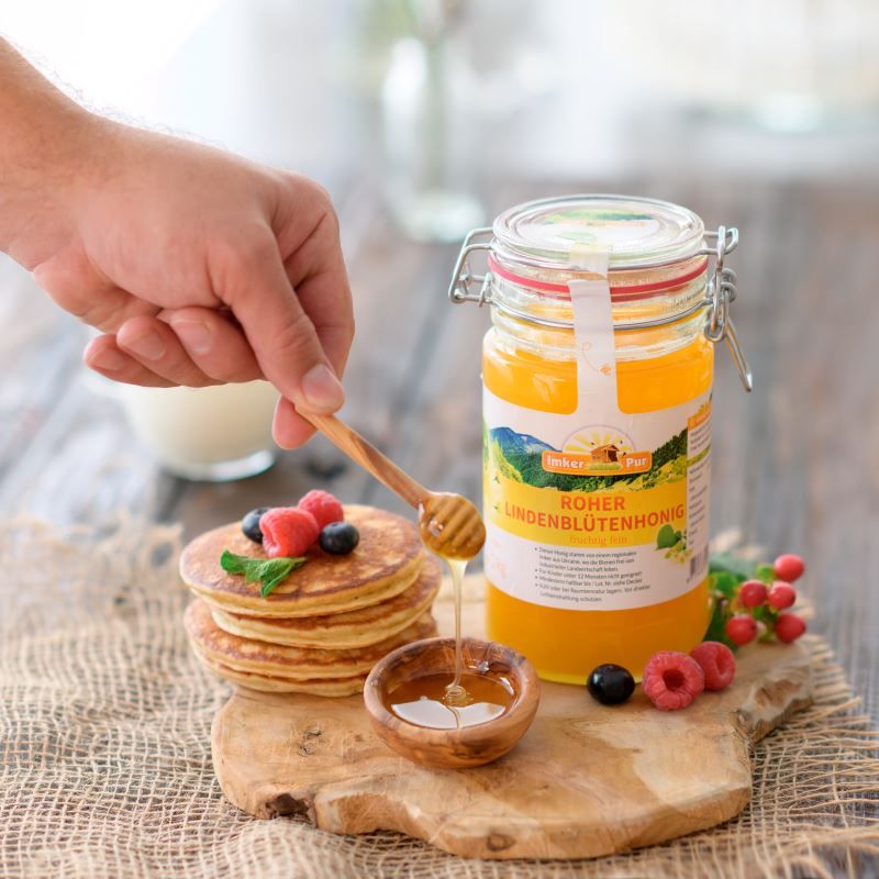 Honigglas mit Etikett, Holzschale mit Honig, Löffel, Pfannkuchen, Beeren. Hand hält Löffel über Honig.