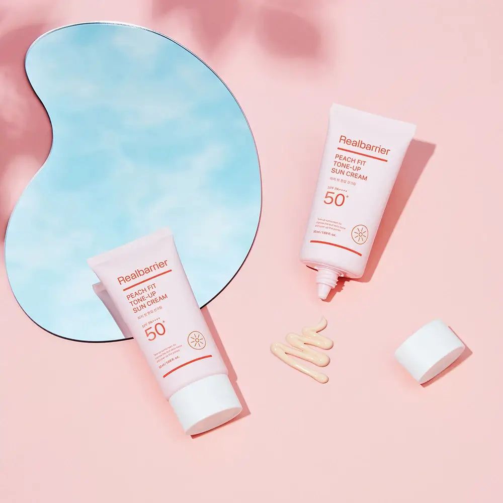 Zwei Tuben, eine geöffnet, Produkt daneben. Spiegel und Produkt auf rosa Untergrund. Aufschrift: Realbarrier Peach Fit Tone-Up Sun Cream 50+.