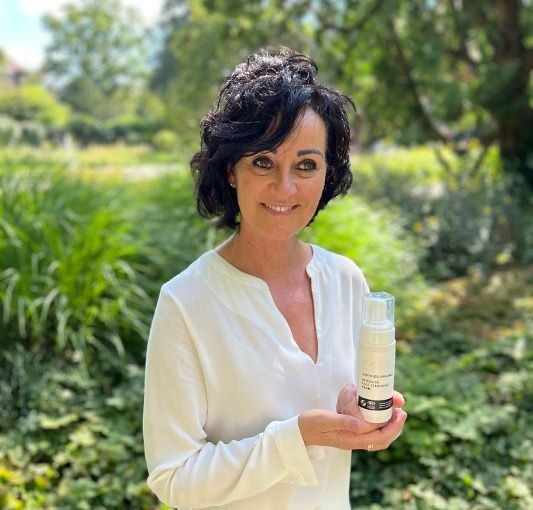 Frau hält Produkt in der Hand. Weißer Spender mit Text und Logo. Grünflächen im Hintergrund.