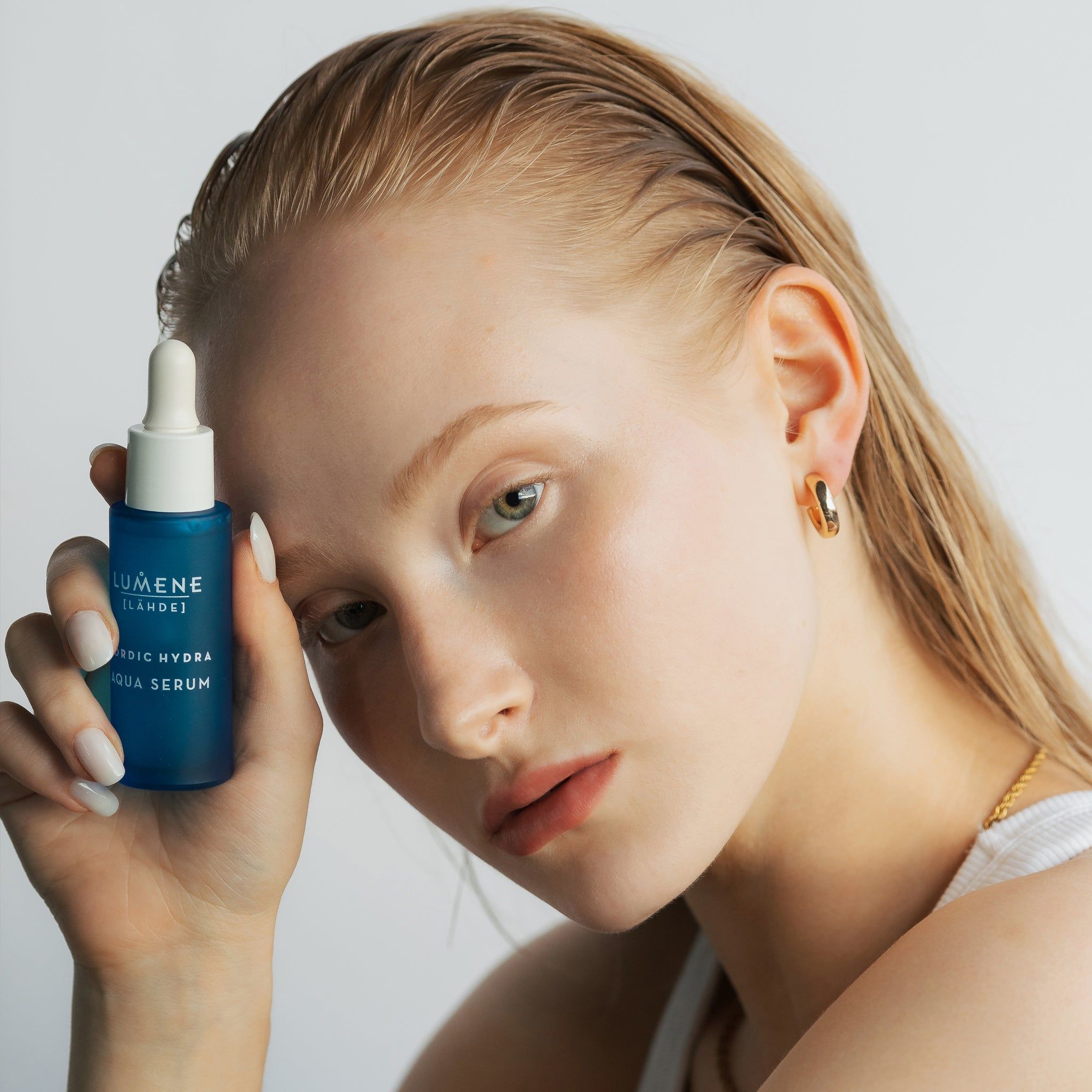 Frau mit Serumflasche. Aufschrift: Lumene [LAHDE] Nordic Hydra Aqua Serum. Frau trägt goldene Ohrringe.