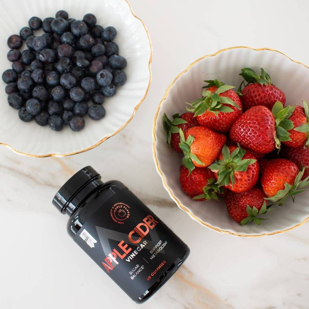 Schwarze Flasche "Apple Cider Vinegar" neben Schalen mit Blaubeeren und Erdbeeren.