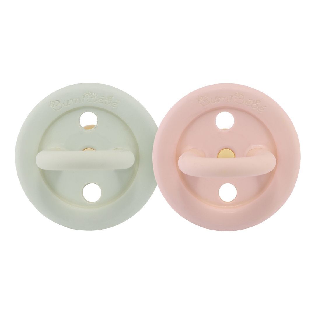 Zwei runde Schnuller, rosa und mintgrün. Mit Griff und Löchern. Aufschrift 'Bumi Bebe'.