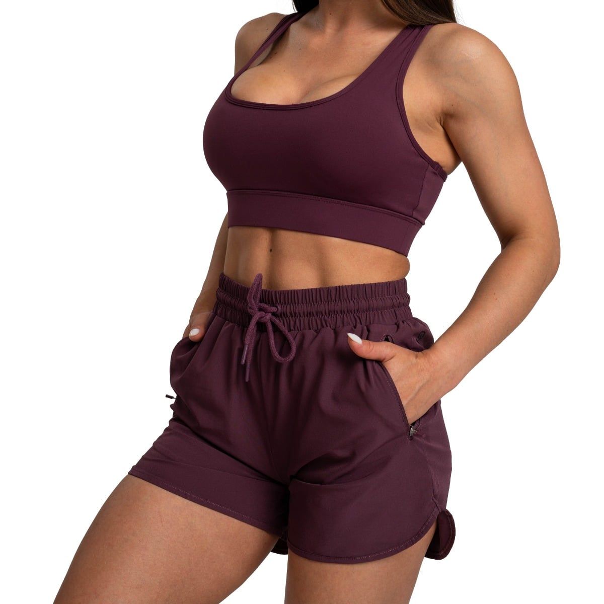 Frau in Aubergine Sport-BH und Shorts mit Kordelzug, Hände in den Taschen.