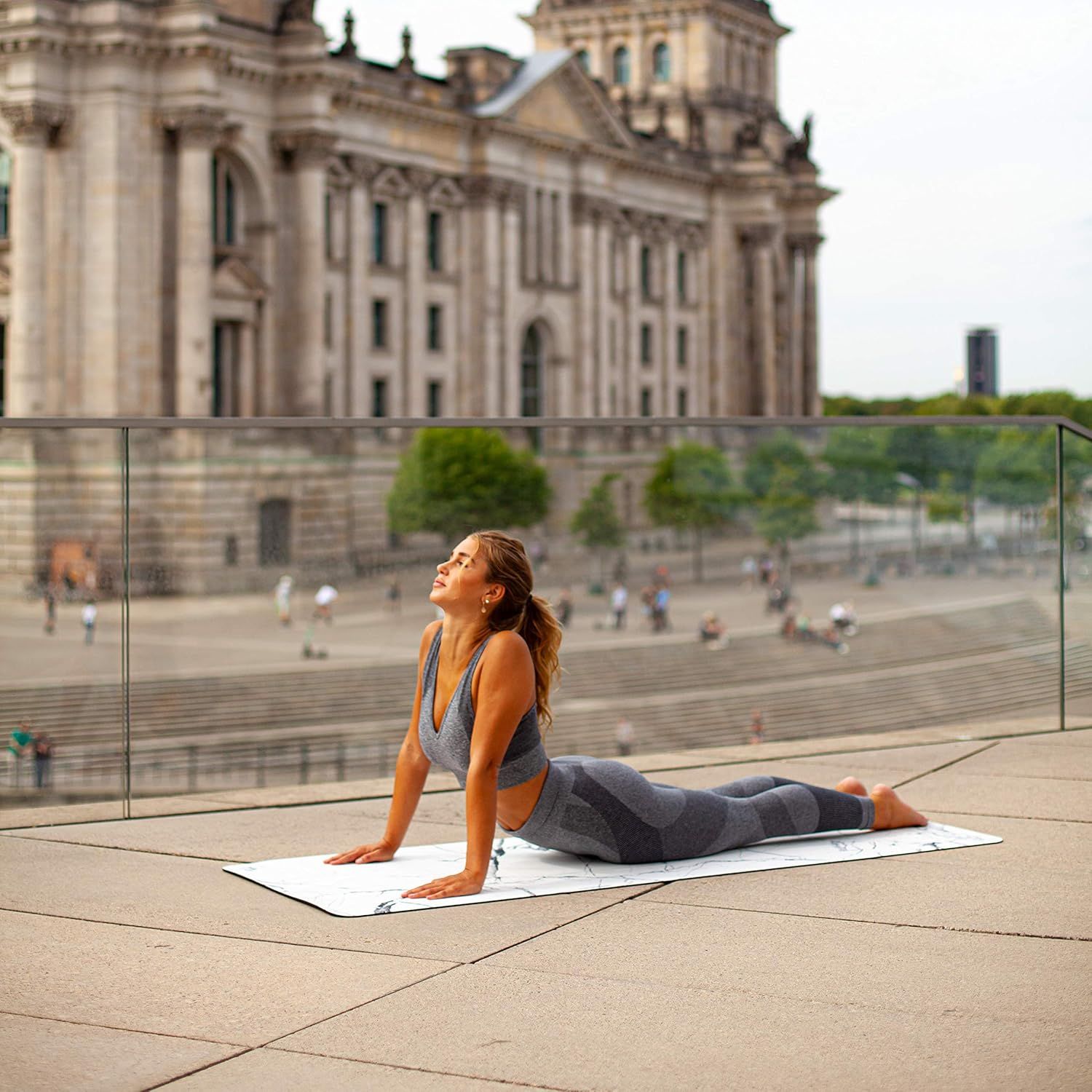 Frau praktiziert Yoga auf einer Yogamatte mit Marmormuster im Freien. Hintergrund: Gebäude.