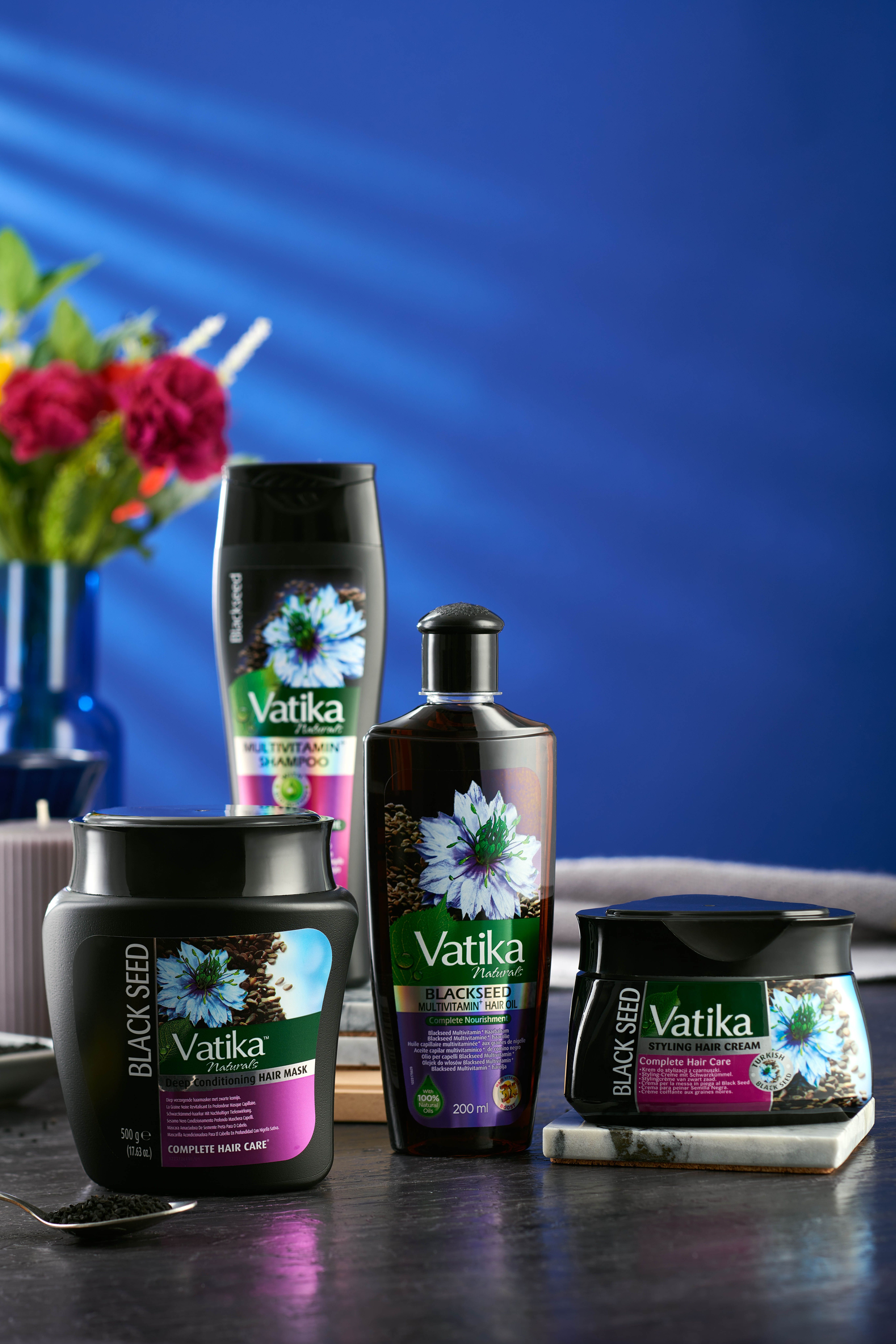 Verschiedene Vatika Naturals Schwarzkümmel Produkte. Flaschen und Tiegel auf blauem Hintergrund, mit Blumen.