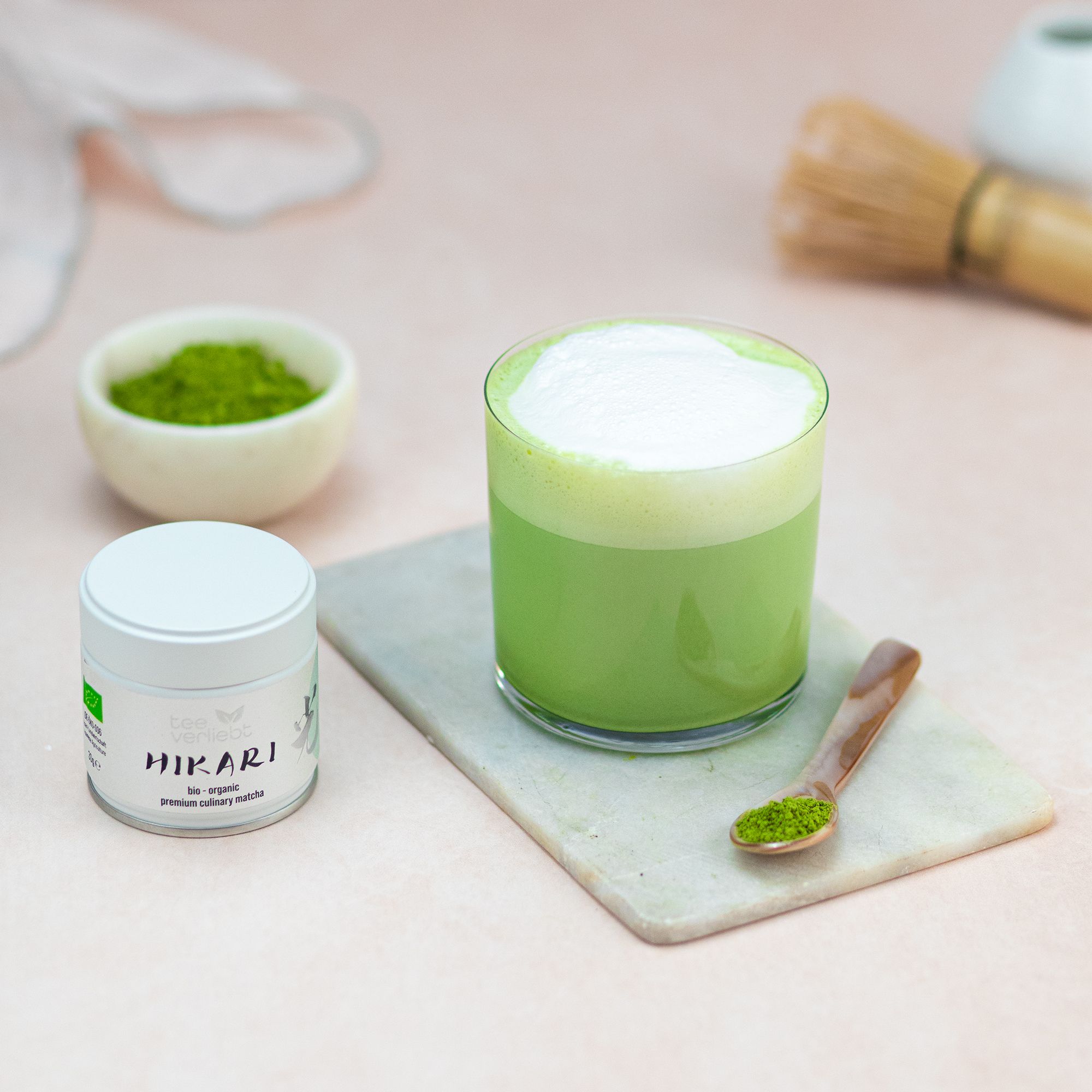 Glas Matcha-Getränk mit Schaum. Dose "teeverliebt Hikari" daneben. Schale mit Matcha-Pulver. Löffel mit Matcha. Marmorplatte.