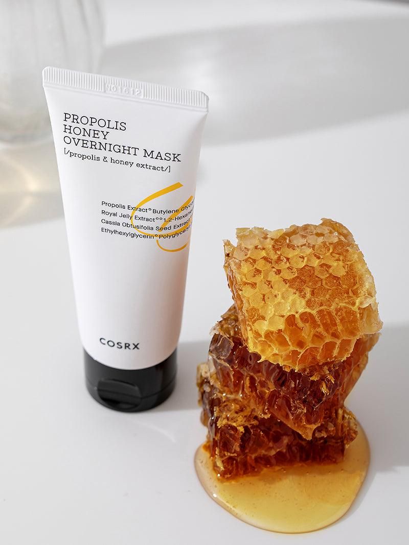Weiße Tube mit schwarzem Deckel neben Honigwabe und Honig. Aufschrift: PROPOLIS HONEY OVERNIGHT MASK. Marke: COSRX.