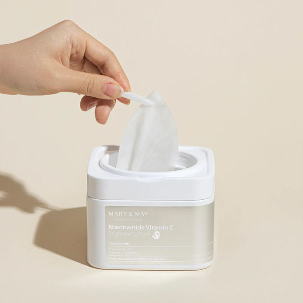 Hand entnimmt eine Maske aus einem weißen Behälter mit MARY & MAY Niacinamide Vitamin C Brightening Mask.