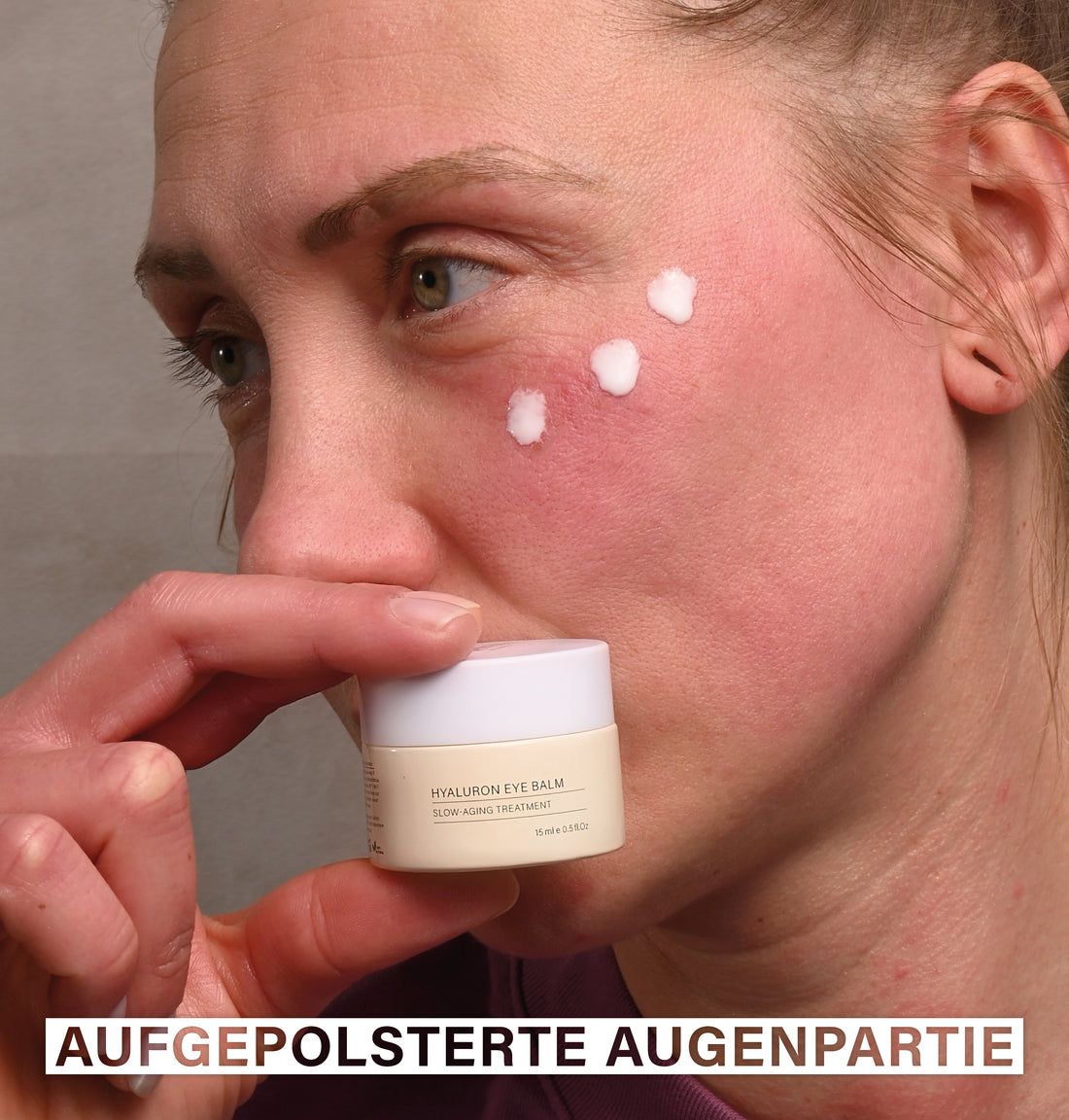 Person hält ein kleines, beiges Tiegel mit weißem Deckel. Auf dem Gesicht sind drei Tupfer Creme aufgetragen.