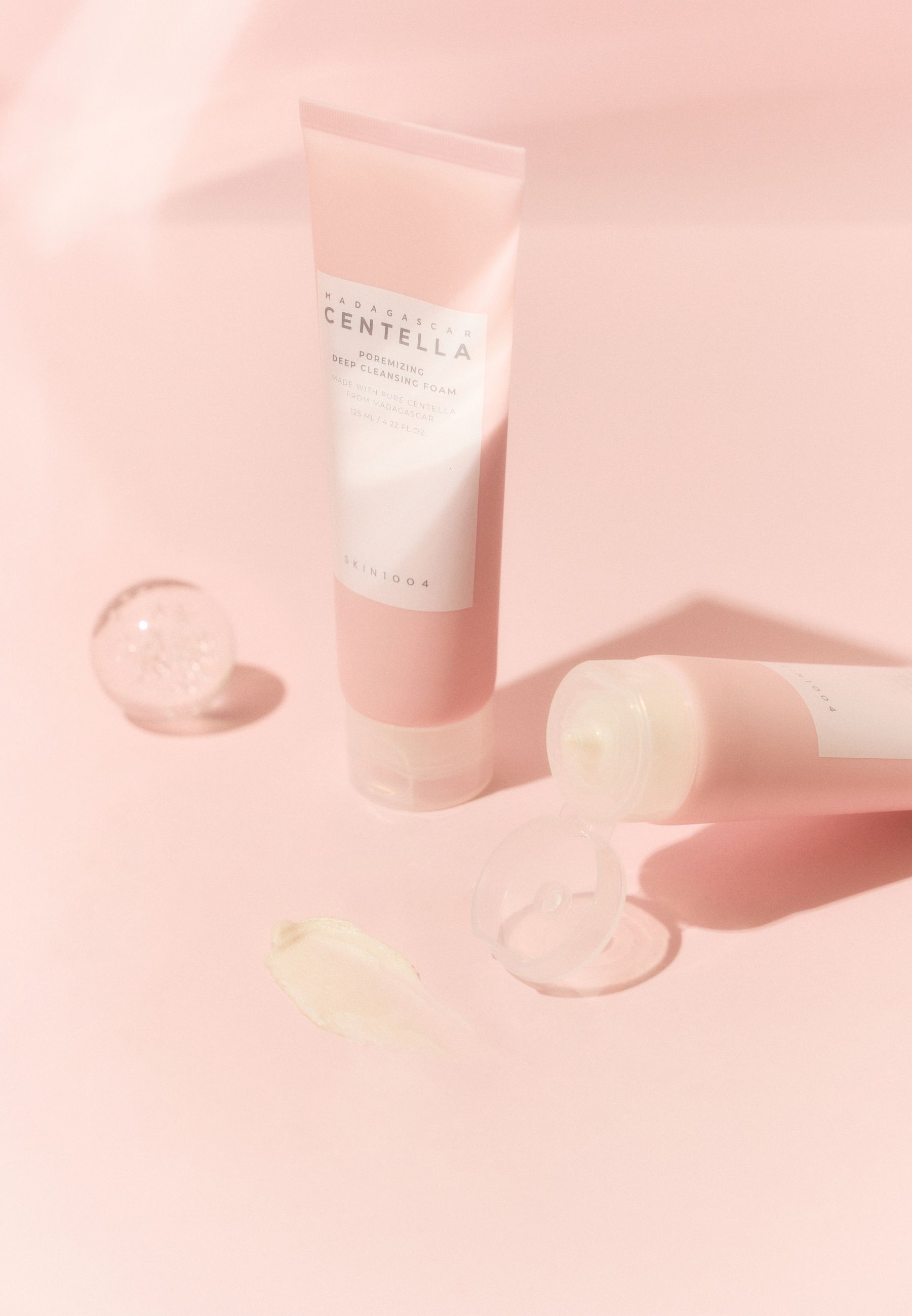 Rosa Tube mit Produktnamen und Logo. Zwei Tuben und eine Kugel auf rosa Oberfläche.