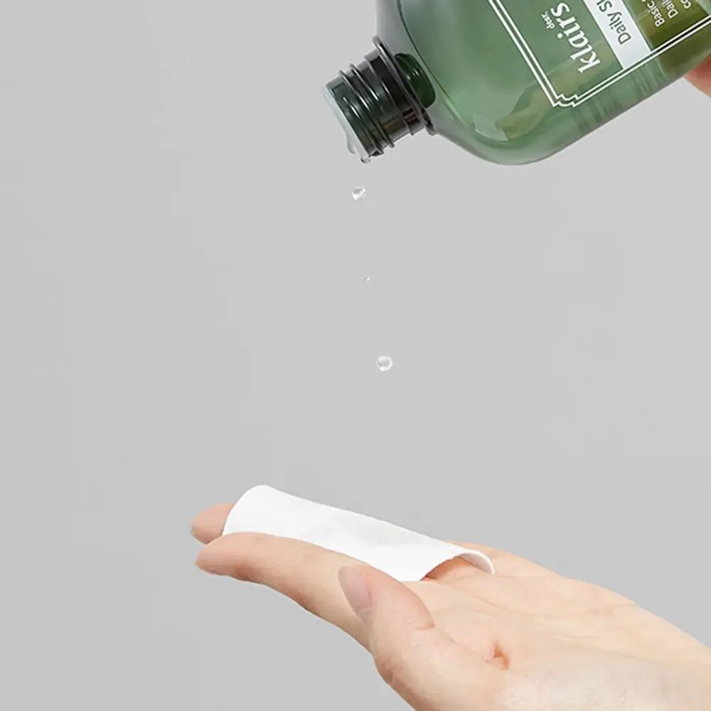 Grüne Flasche gießt Flüssigkeit auf Wattepad. Hand im Bild. Helle Umgebung.