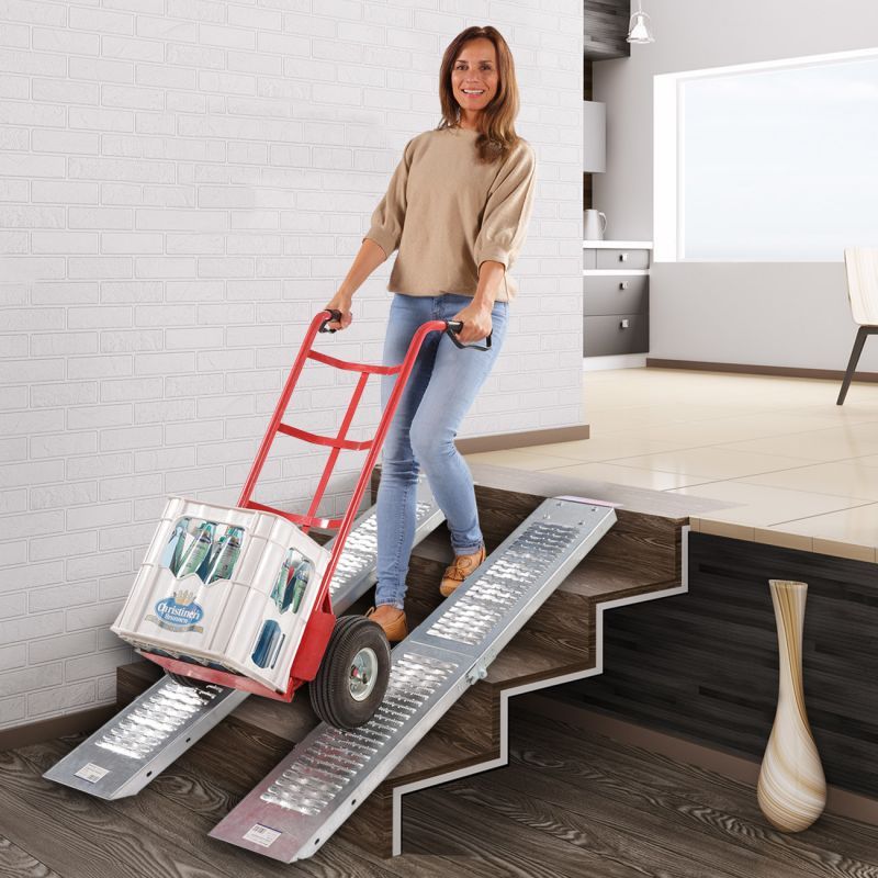 Frau benutzt zwei silberne Auffahrrampen, um eine Kiste mit einer Sackkarre über Treppen zu transportieren.