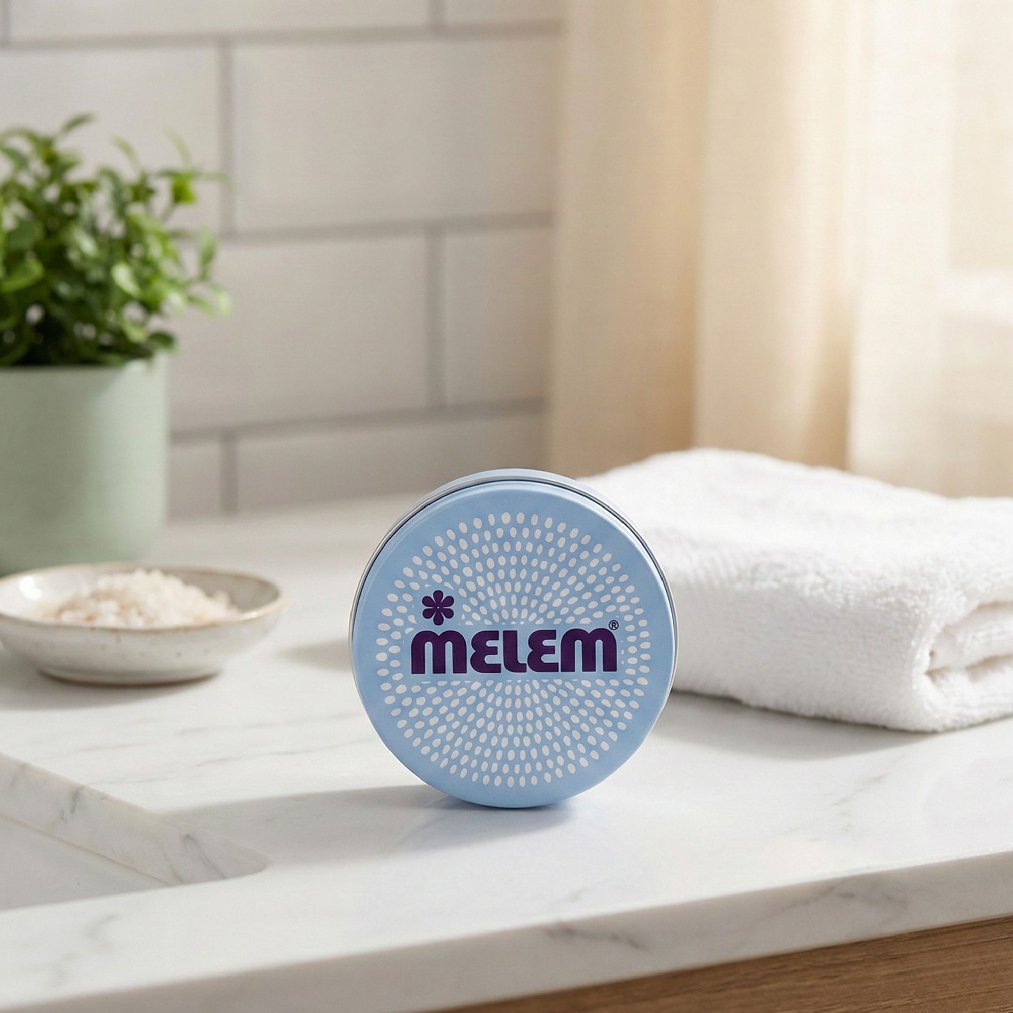 Hellblaues, rundes Produkt mit dem lila Schriftzug "MELEM". Auf einer weißen Oberfläche, neben einem Handtuch und einer Pflanze.