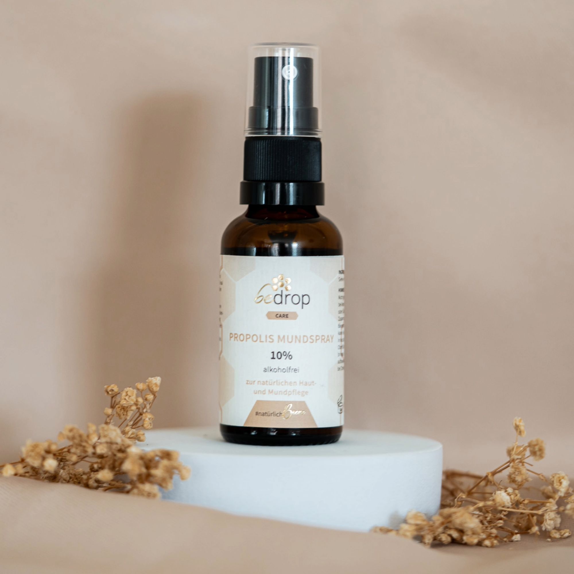 Braune Glasflasche mit schwarzem Sprühkopf. Etikett mit Produktbezeichnung und Logo. Auf weißem Podest, umgeben von Dekoration.