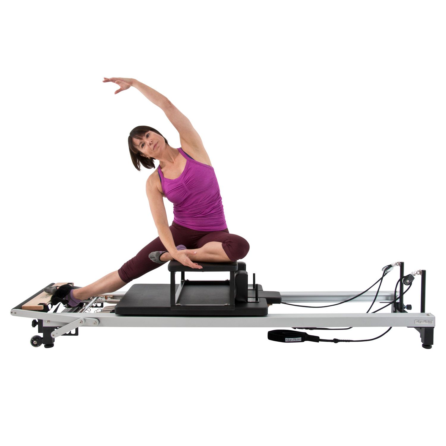 Frau sitzt auf einer schwarzen Pilates-Sitzbox auf einem Reformer. Sie dehnt sich, Arm oben. Der Reformer ist silberfarben.
