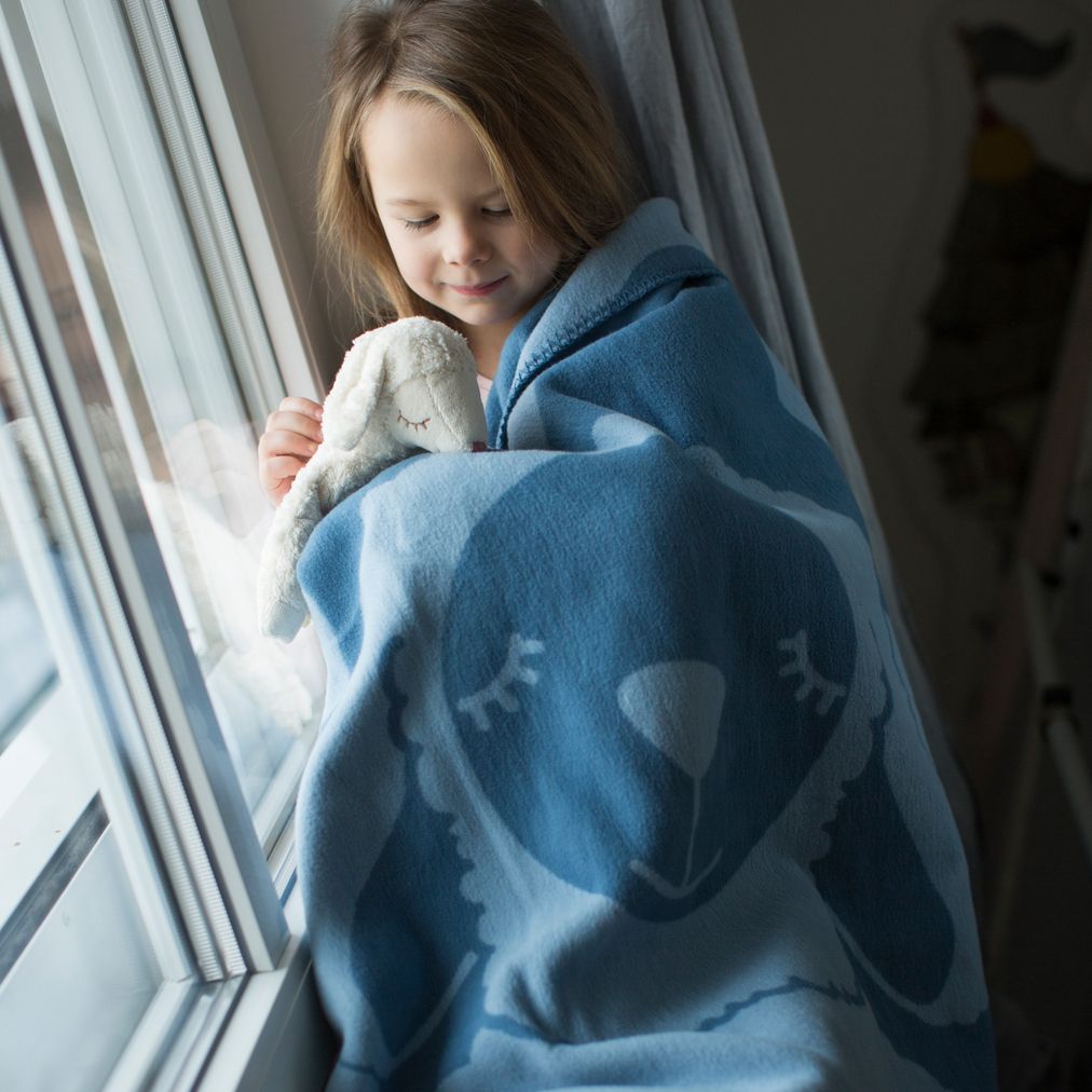 Blaues Plaid mit Schafmotiv, um ein Kind gewickelt. Kind hält Stofftier. Fenster im Hintergrund.