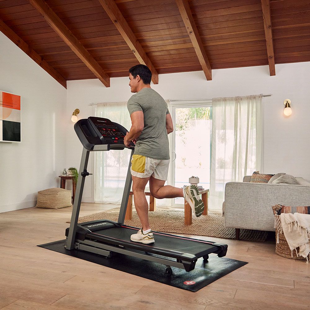 Mann läuft auf einem Laufband. Schwarze Lauffläche, grauer Rahmen. Konsole. Schwinn-Logo. In Wohnraum.