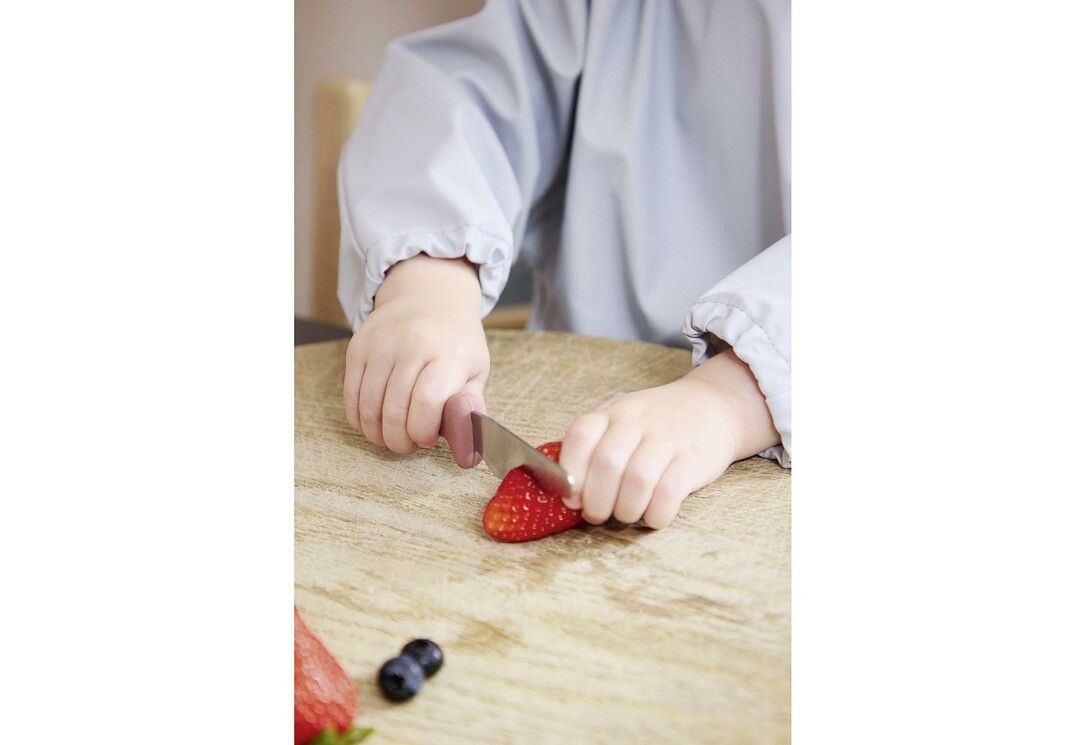 Kind schneidet Erdbeere mit rosa FILIBABBA Kindermesser. Holzbrett, Erdbeere und Blaubeeren liegen daneben. Kind trägt Schürze.