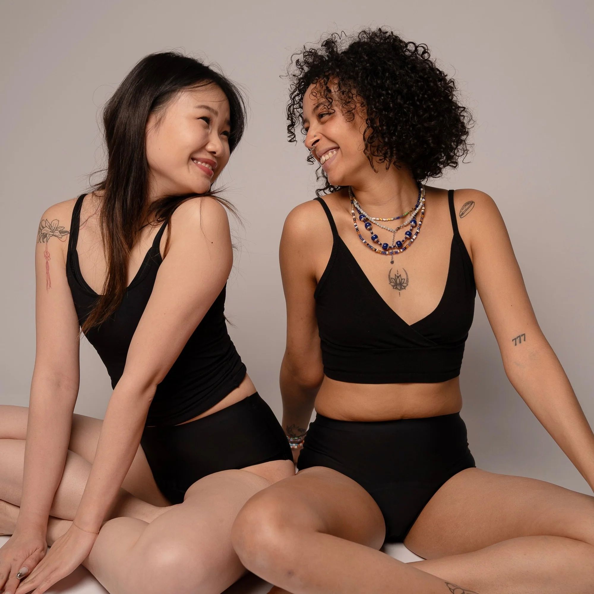 Zwei Frauen sitzen. Beide tragen schwarze Slips und Tanktops. Lächeln sich an.