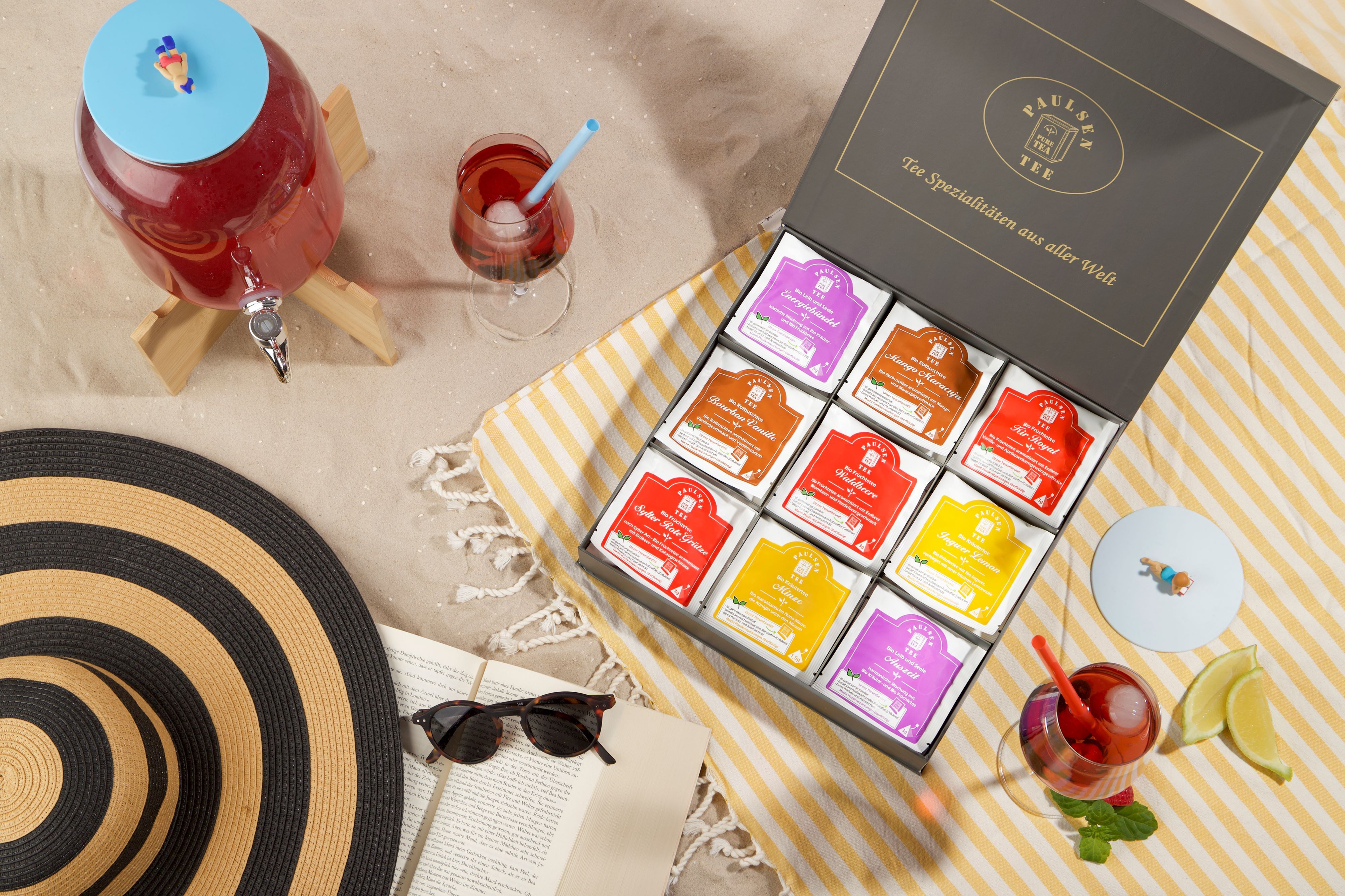 Geöffnete Geschenkbox mit Teebeuteln, neben Getränken und Strandutensilien. Aufschrift: Paulsen Tee.