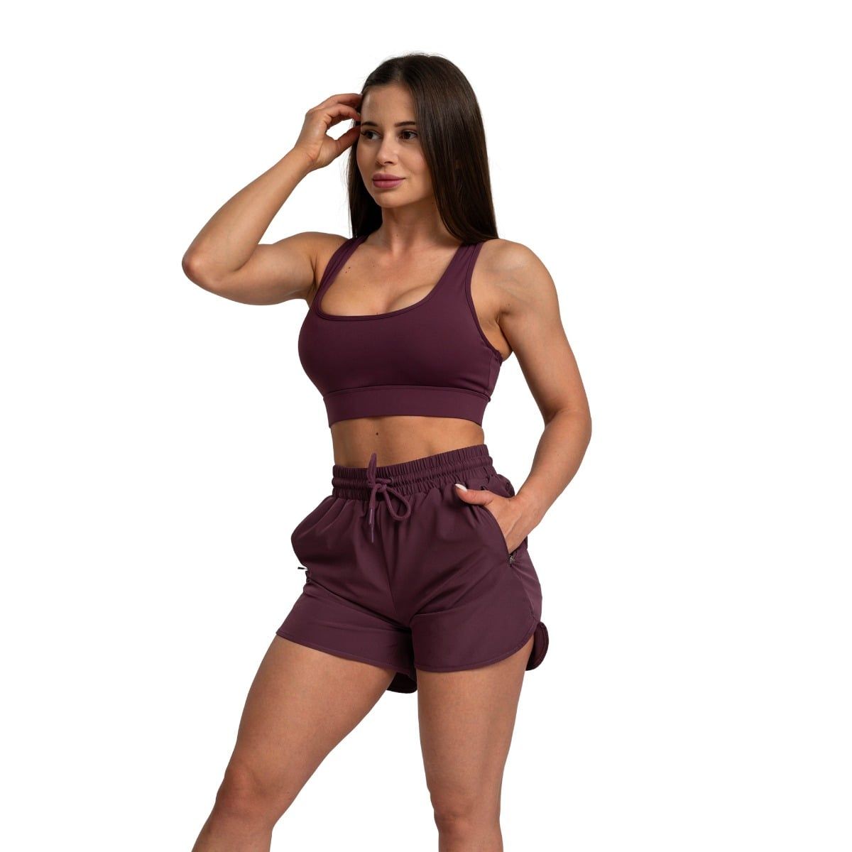 Frau in Aubergine Sport-BH und Shorts mit Kordelzug, eine Hand in der Tasche.