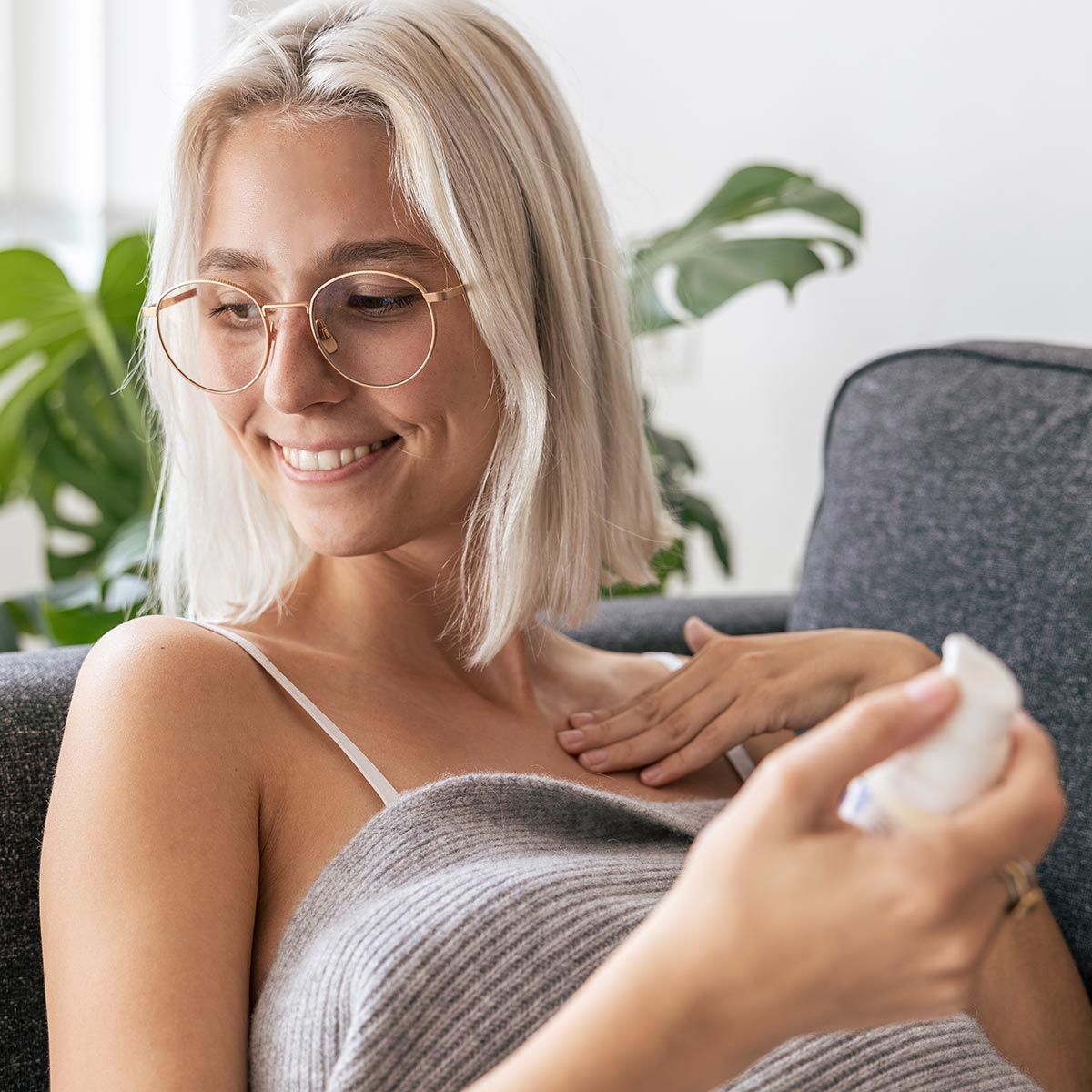 Person massiert Balsam auf die Brust. Balsam-Spender in der Hand. Person trägt Brille und lächelt. Graue Decke.