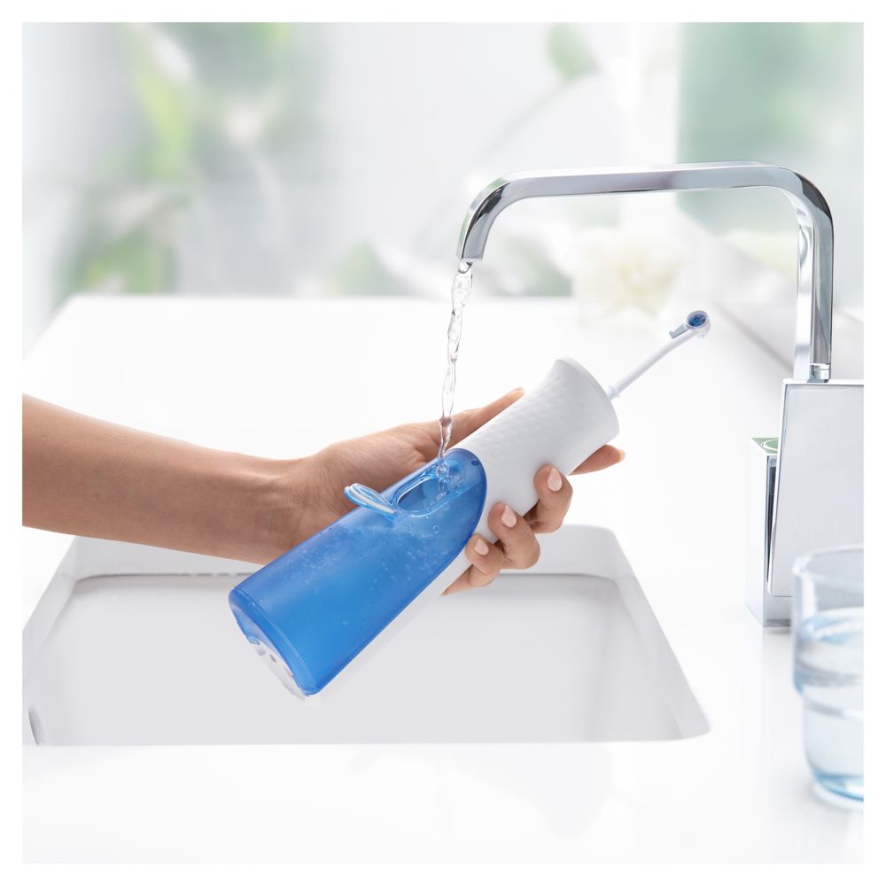 Person hält Munddusche unter fließendem Wasser. Blauer Wassertank sichtbar. Waschbecken im Hintergrund.