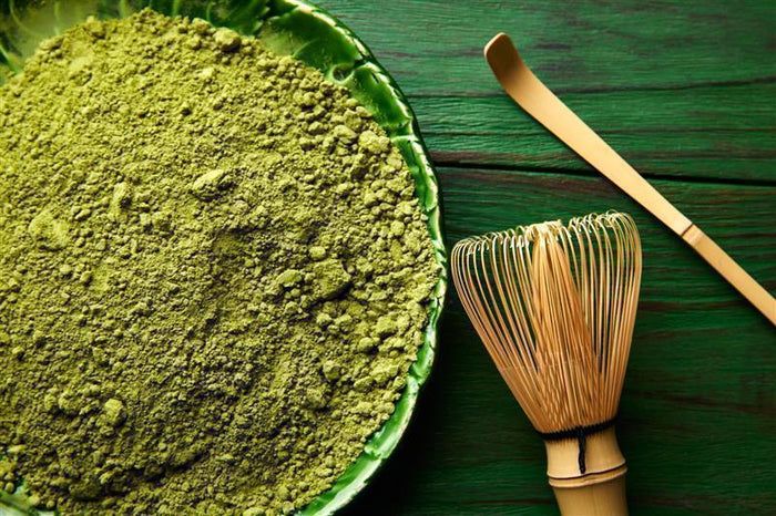 Grünes Matcha-Pulver in einer Schale. Daneben ein Bambus-Besen und ein Löffel. Auf grünem Holzuntergrund.