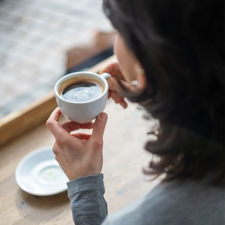 Person hält eine weiße Tasse mit Kaffee. Auf einem Holztisch. Untertasse daneben.