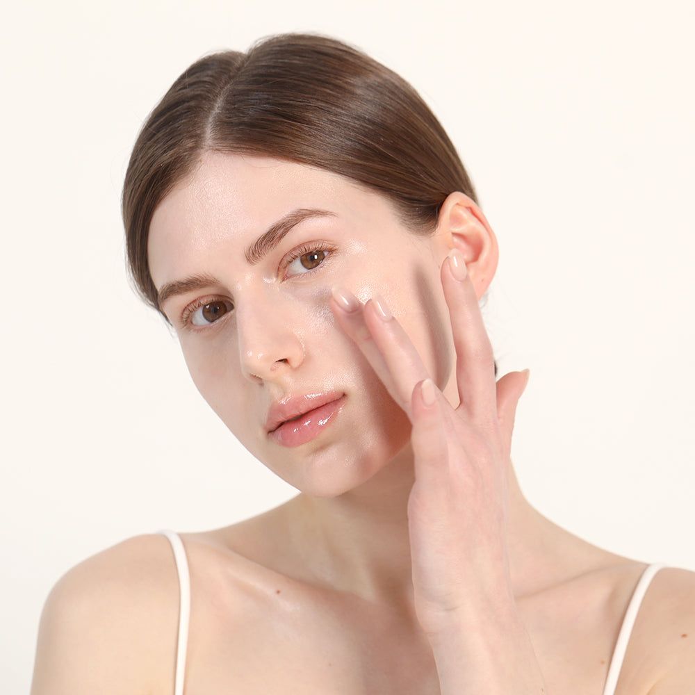 Frau trägt Serum auf das Gesicht auf. Helle Haut, weißes Top. Produkt nicht sichtbar.