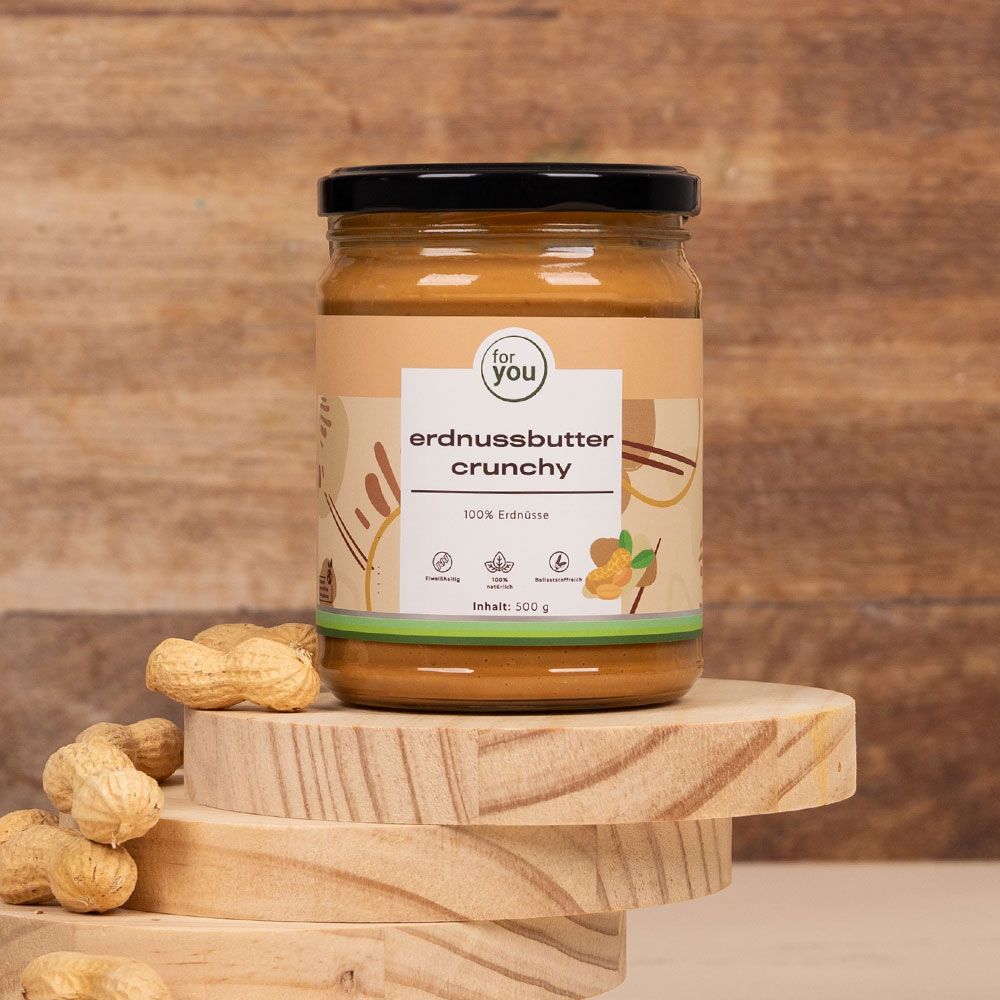 Glas Erdnussbutter Crunchy auf Holzscheiben. Daneben Erdnüsse. Aufschrift: for you, erdnussbutter crunchy, 100% Erdnüsse. Inhalt: 500 g.