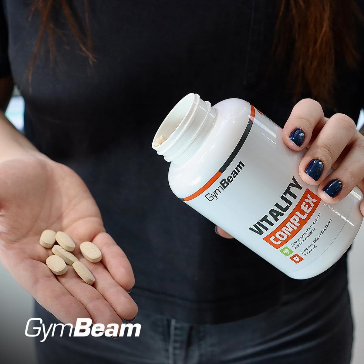 Eine Hand hält GymBeam Vitality Complex Tabletten, daneben eine offene Flasche.