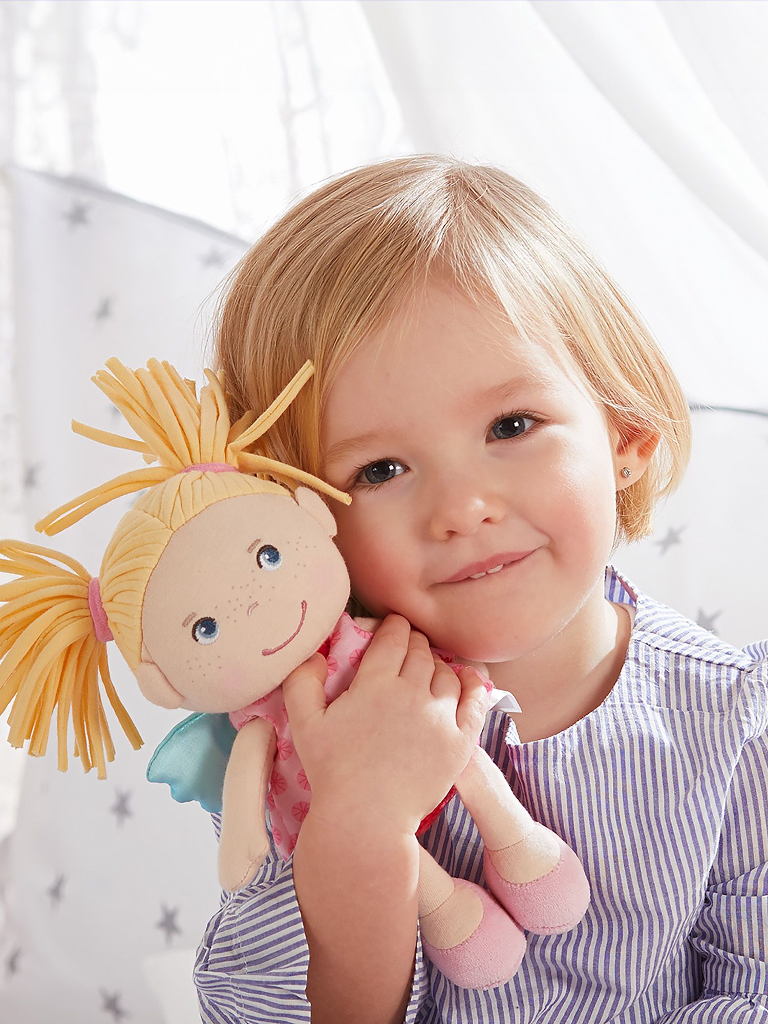 Kleinkind hält Stoffpuppe Finja. Kind lächelt. Puppe mit blonden Zöpfen, rosa Kleid mit Blumenmuster, Flügeln.