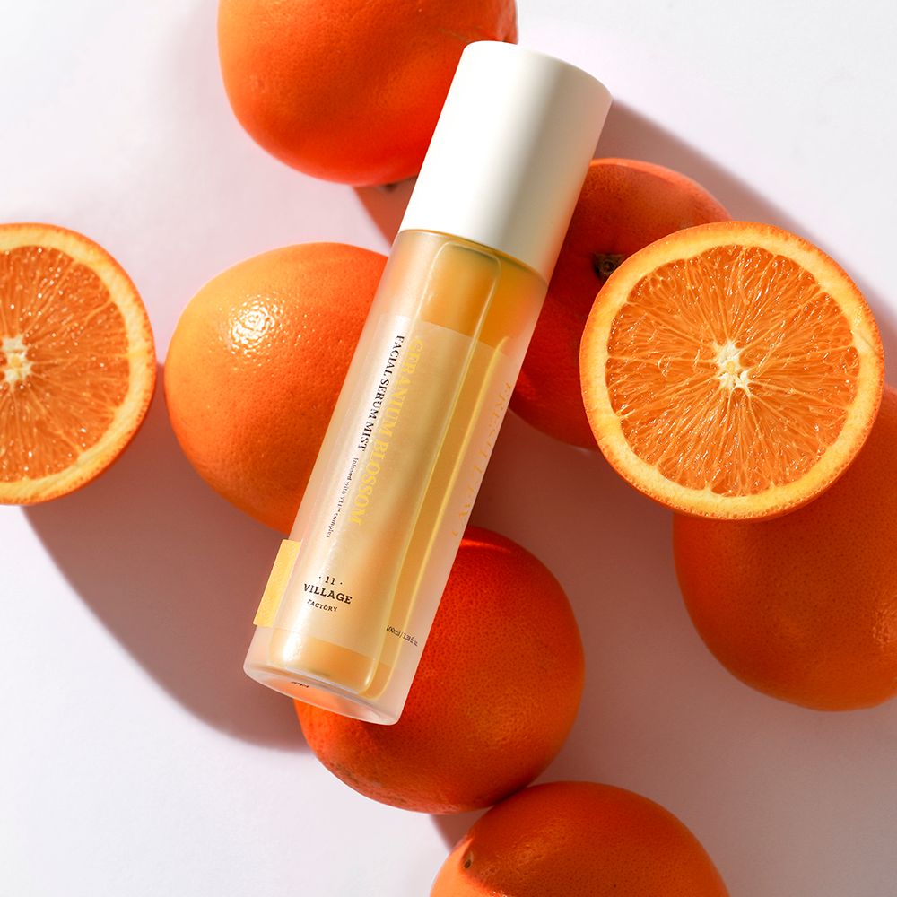 Produktflasche umgeben von Orangen. Text: FACIAL SERUM MIST, -11- VILLAGE FACTORY. Weißer Deckel. Orangen in Scheiben.