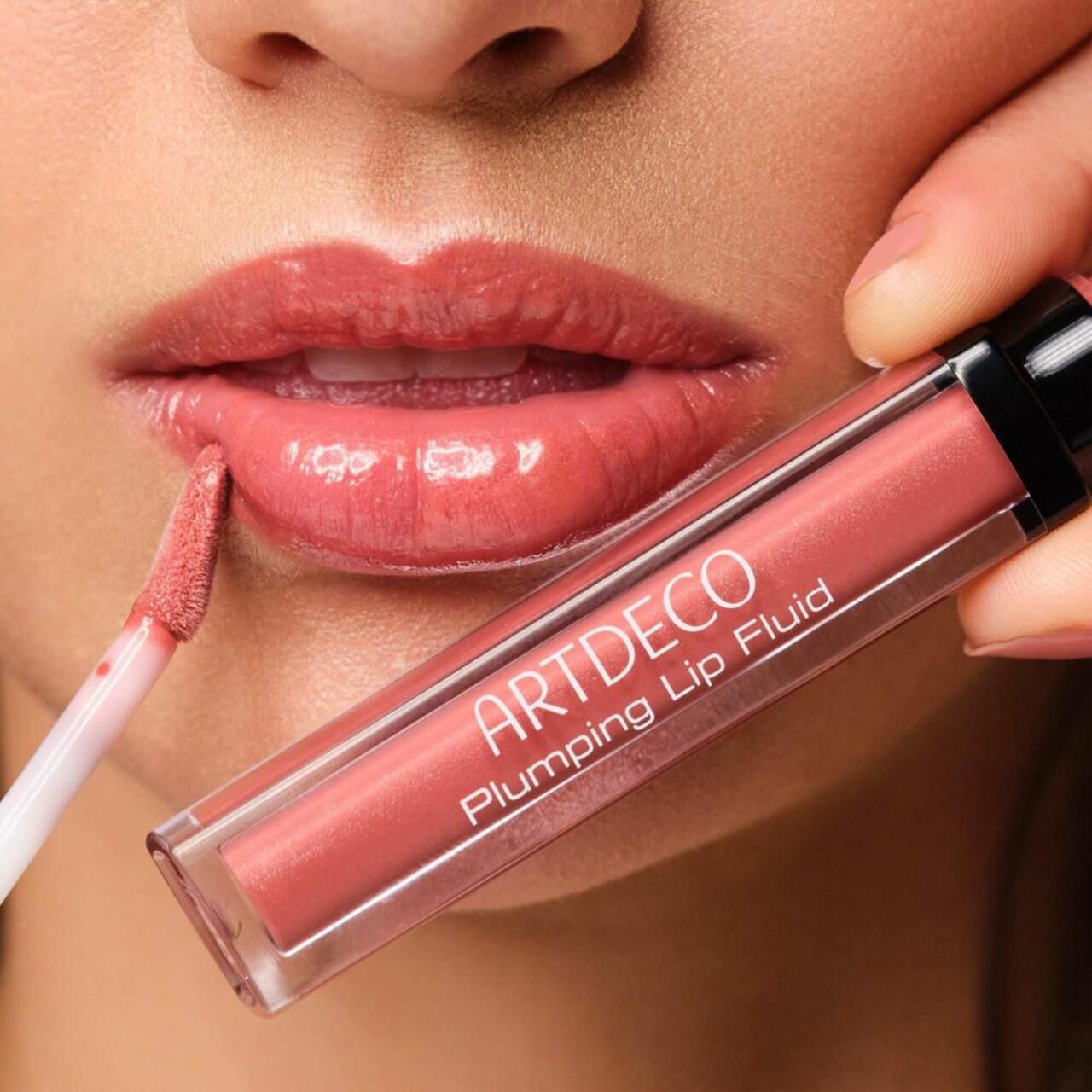 Person trägt Artdeco Plumping Lip Fluid auf. Produkt und Applikator sichtbar. Lippen glänzend, rosafarben.