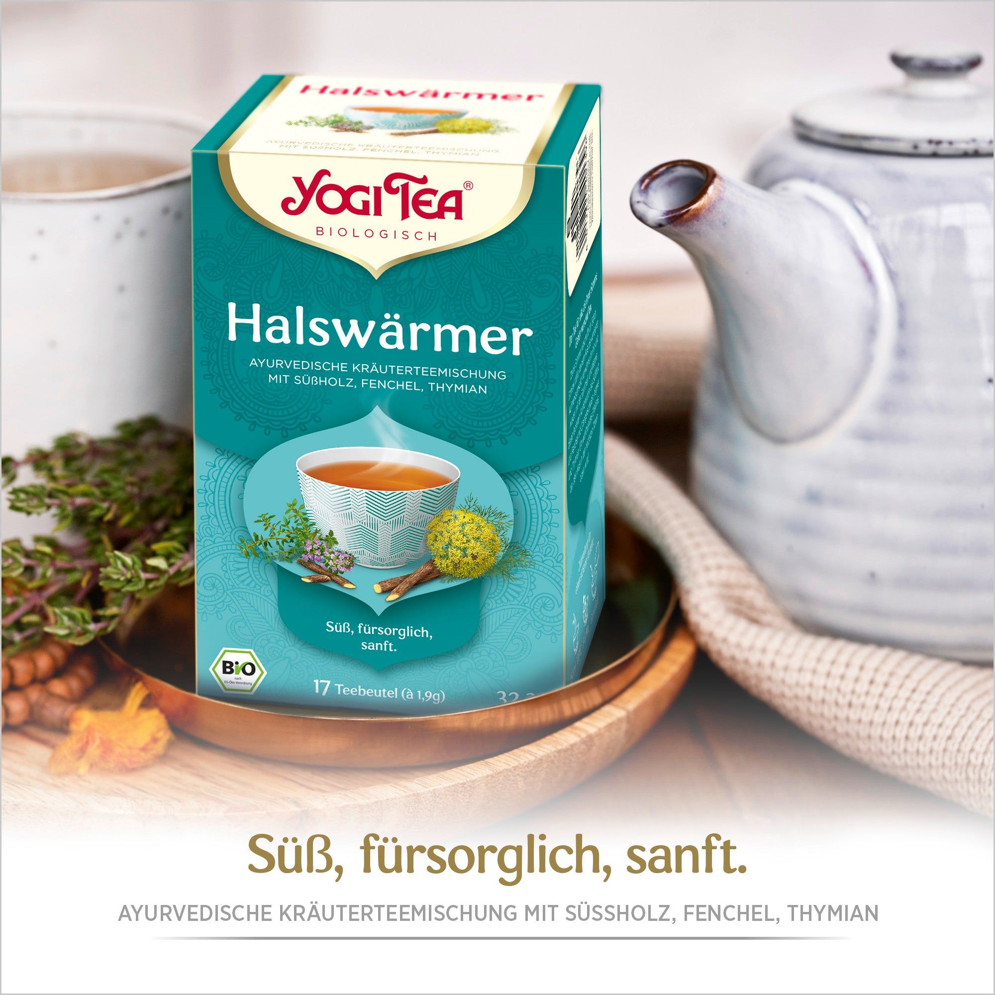 Yogi Tea Halswärmer Packung. Türkis-grüne Schachtel mit Logo, Produktname und Abbildung einer Tasse Tee. Auf Tisch mit Teekanne.