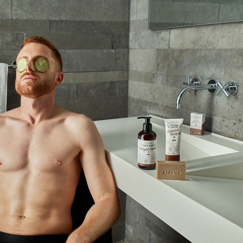 Mann mit Gurkenscheiben auf den Augen, neben Waschbecken mit Monsieur BARBIER Produkten. Seife, Gesichtsreiniger, Feuchtigkeitspflege.