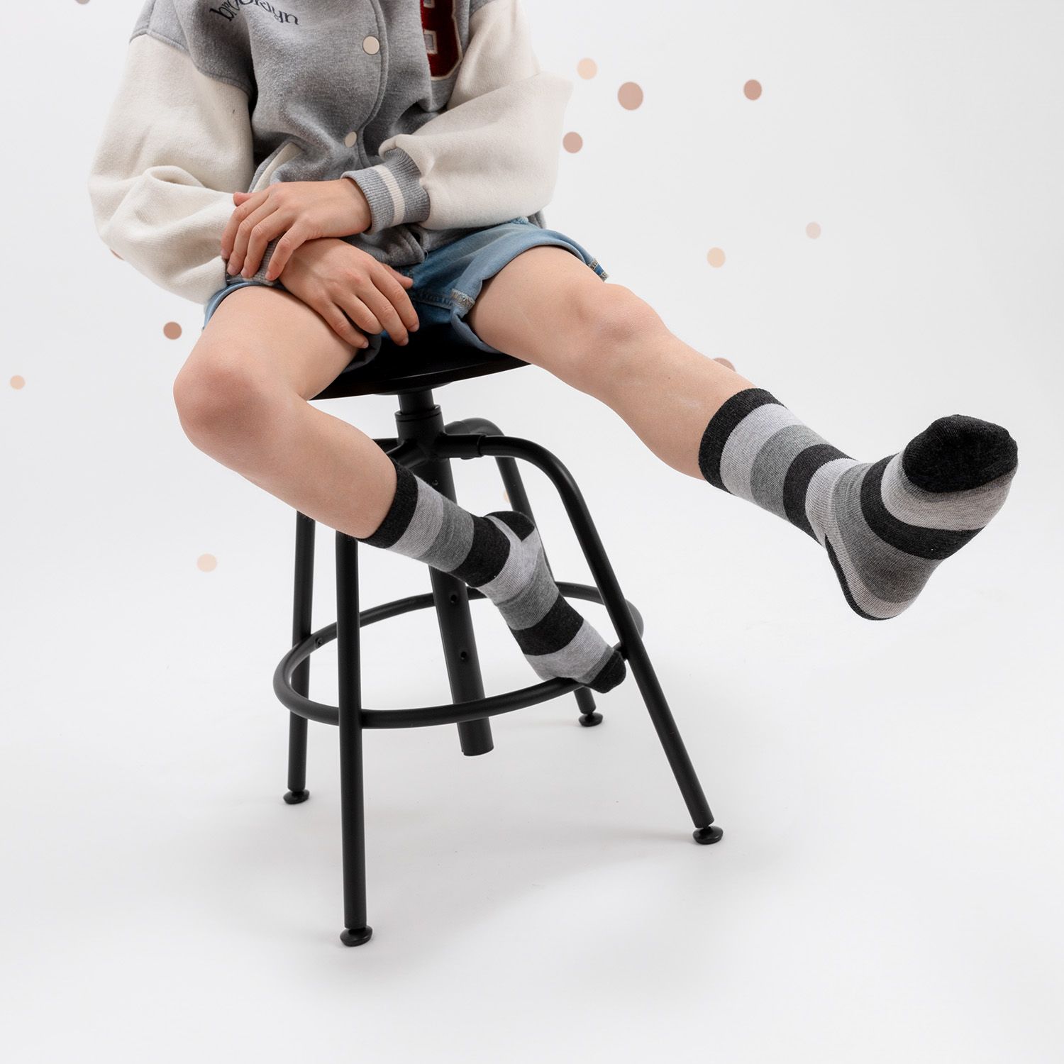 Kind sitzt auf einem Hocker, trägt gestreifte Socken in Grau und Schwarz. Kurze Hose und Jacke.