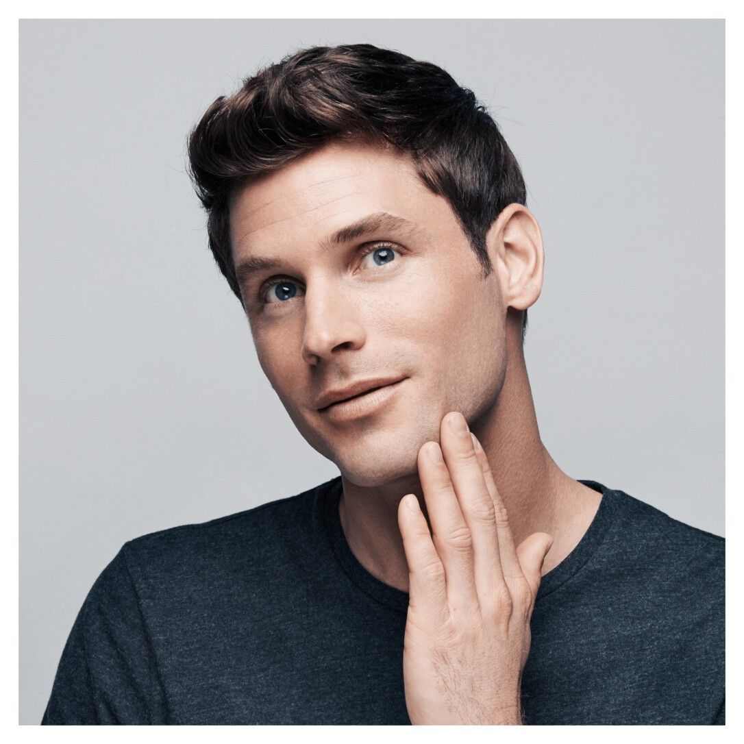 Porträt eines Mannes, der sich mit der Hand am Kinn berührt. Er hat braunes Haar und trägt ein dunkles Hemd.