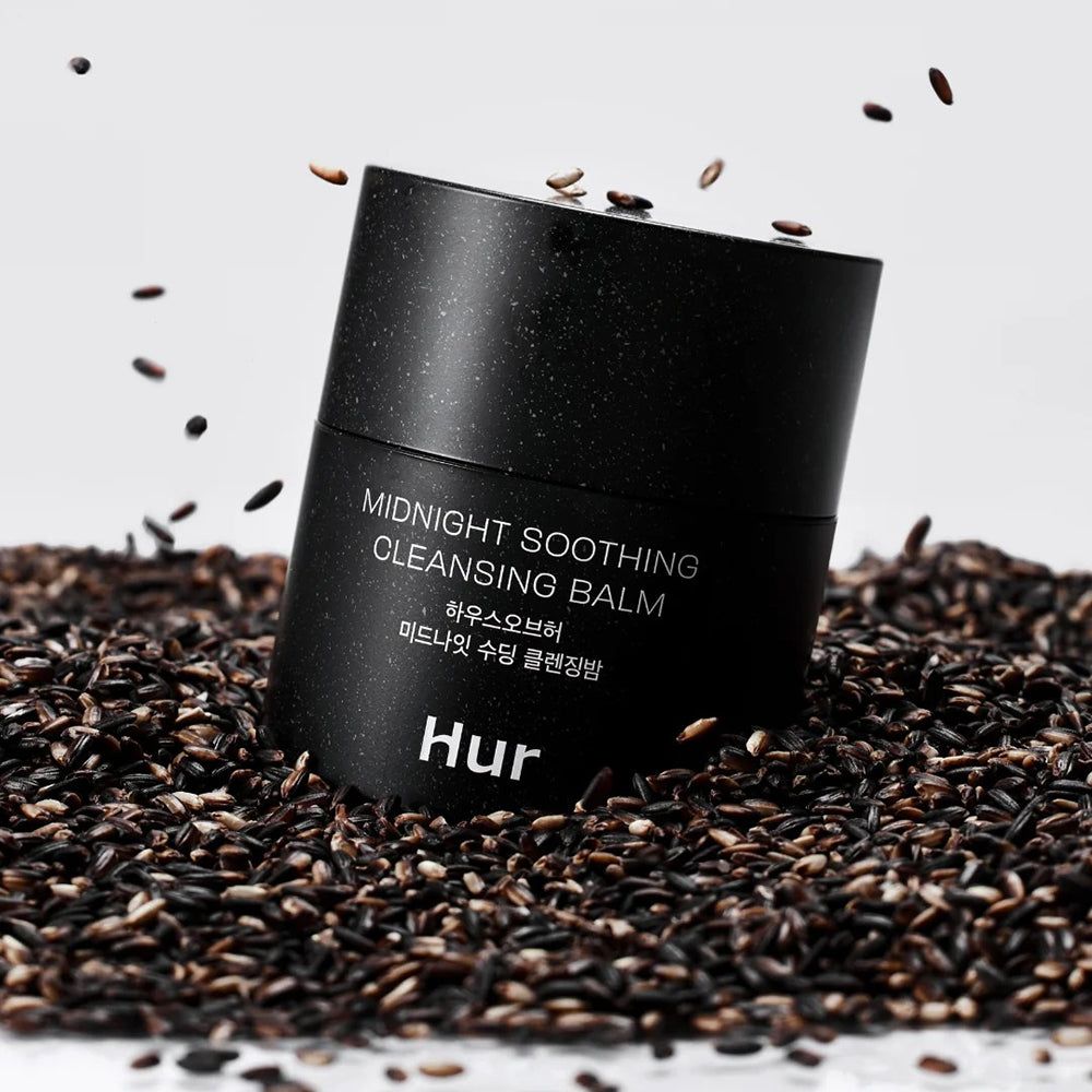 Schwarzer Tiegel mit Aufschrift: Midnight Soothing Cleansing Balm. Marke: Hur. Umgeben von schwarzen Samen. Hintergrund weiß.